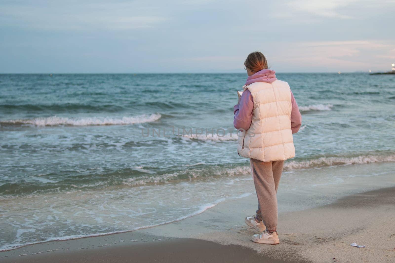 Portrait of an excited girl in her hands with a phone, standing on a beach with a blue sea. Sad and depressed young woman standing alone on the beach by the sea. Depression concept. High quality photo