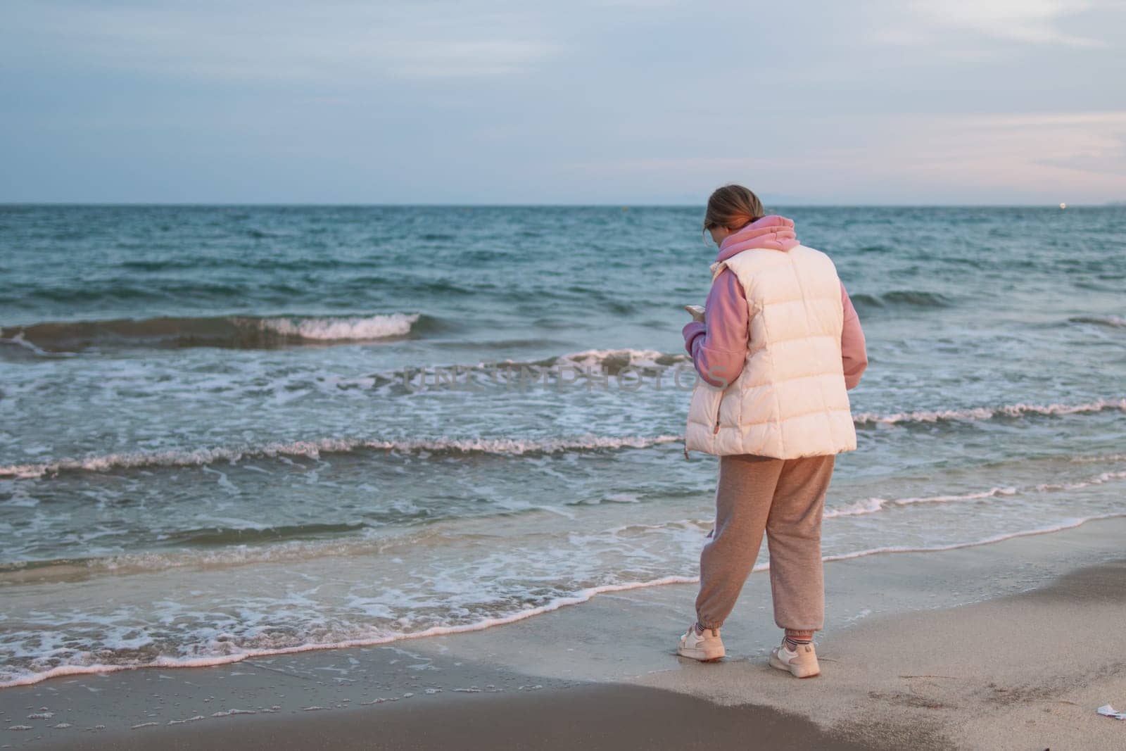 A girl of European make in casual clothes stands near the ocean with a phone in her hands is sad and looks into the phone. by PopOff