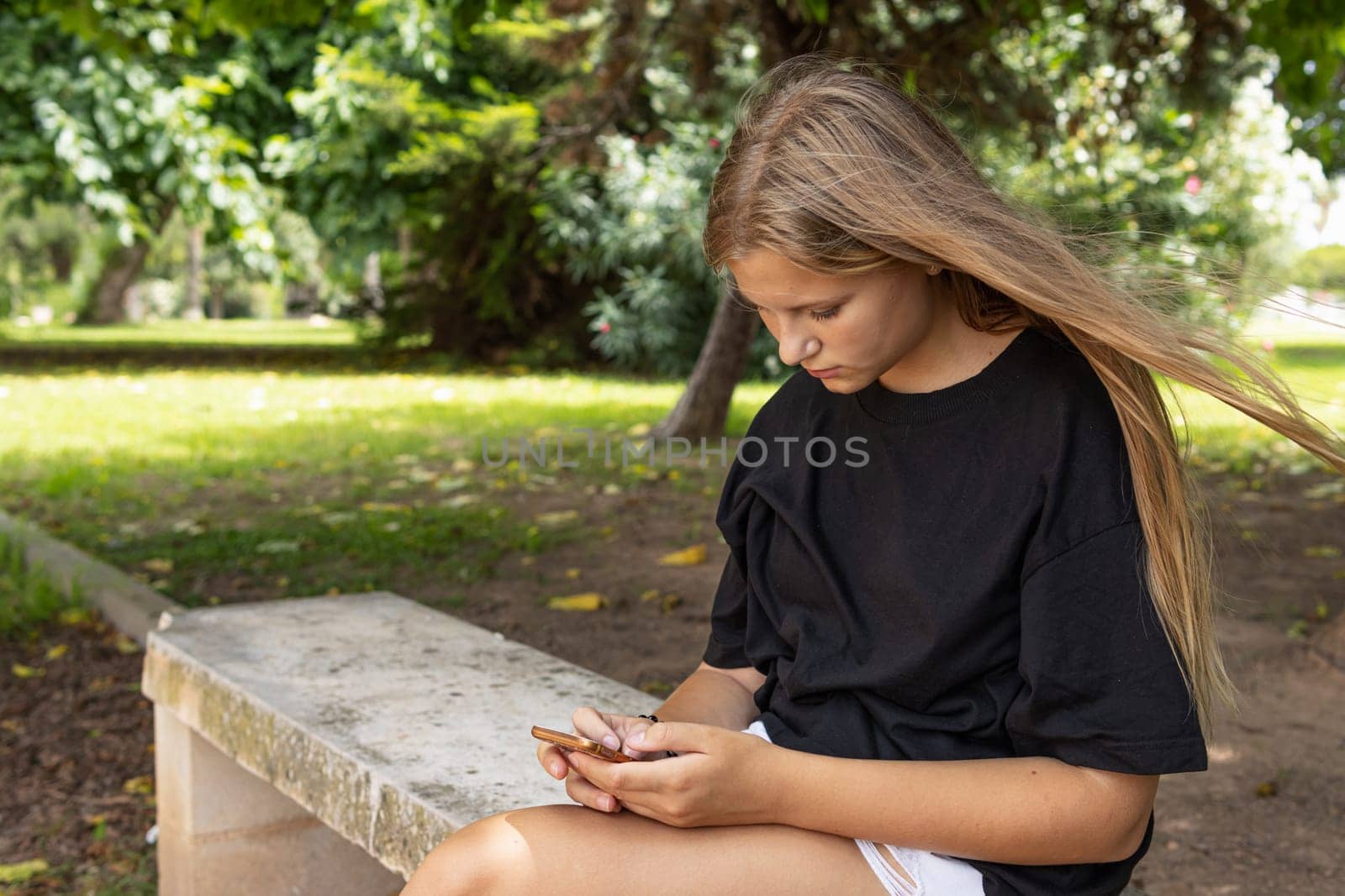 teenage girl in shorts and a black T-shirt sits on a park bench and looks at the phone by PopOff