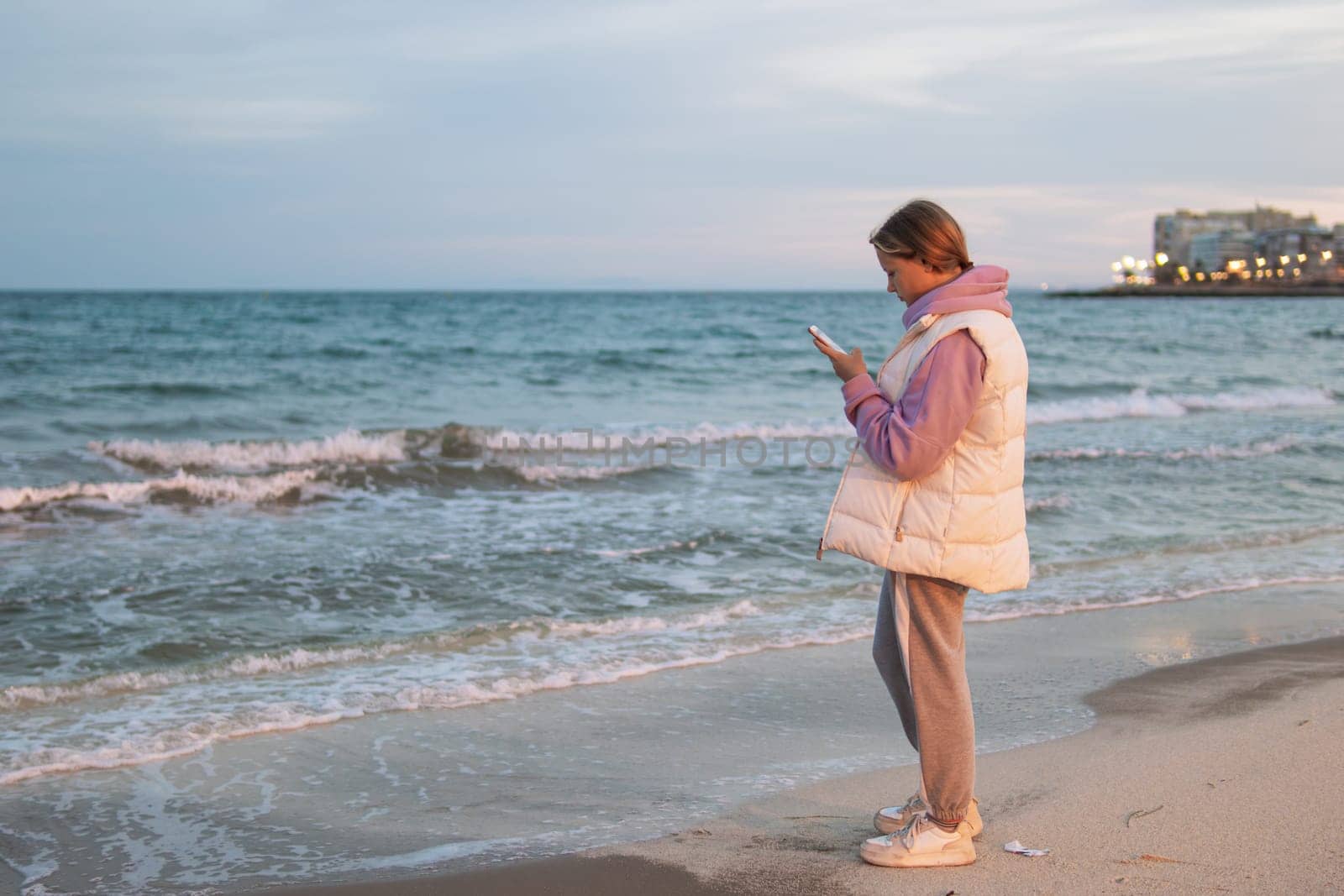 Portrait of an excited girl in her hands with a phone, standing on a beach with a blue sea. Sad and depressed young woman standing alone on the beach by the sea. Depression concept. High quality photo