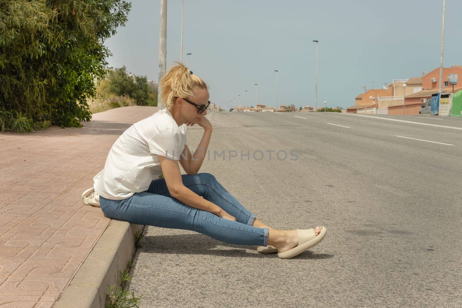 a sad girl sits on the sidewalk in jeans and a T-shirt and glasses, looks at the road, there is a place for an inscription. High quality photo