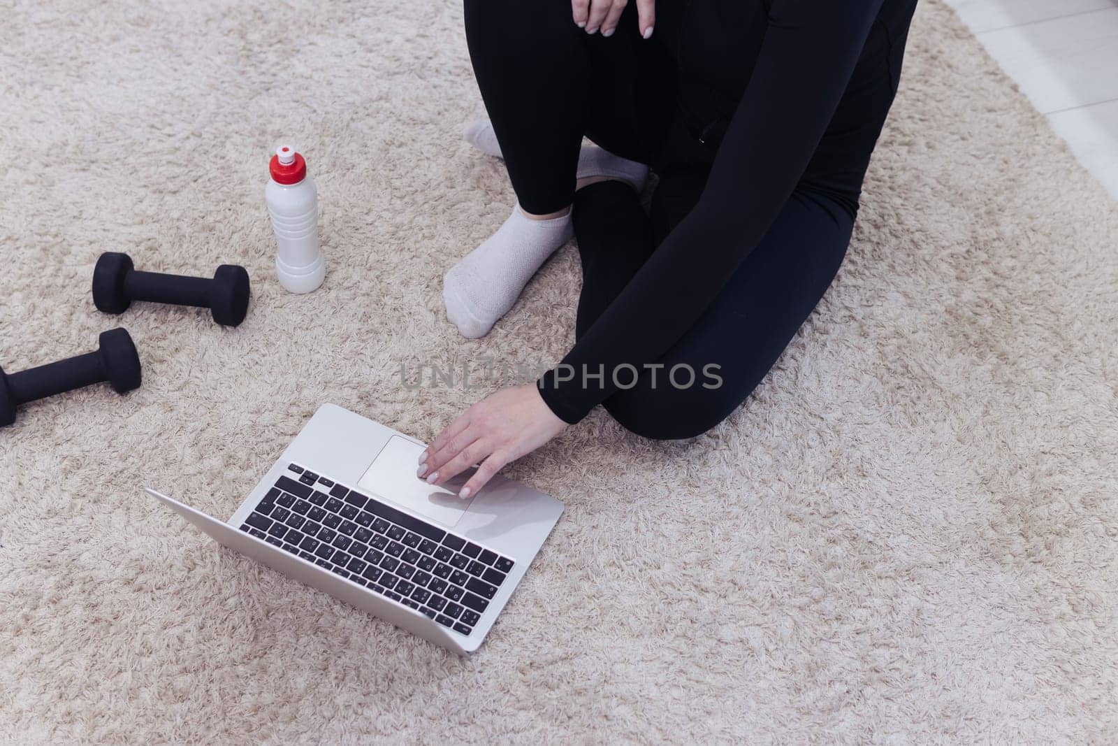 Athletic middle-aged woman in black sportswear sits on the floor on a carpet with dumbbells and a bottle of water and uses a laptop at home in the room. The concept of sports and recreation.