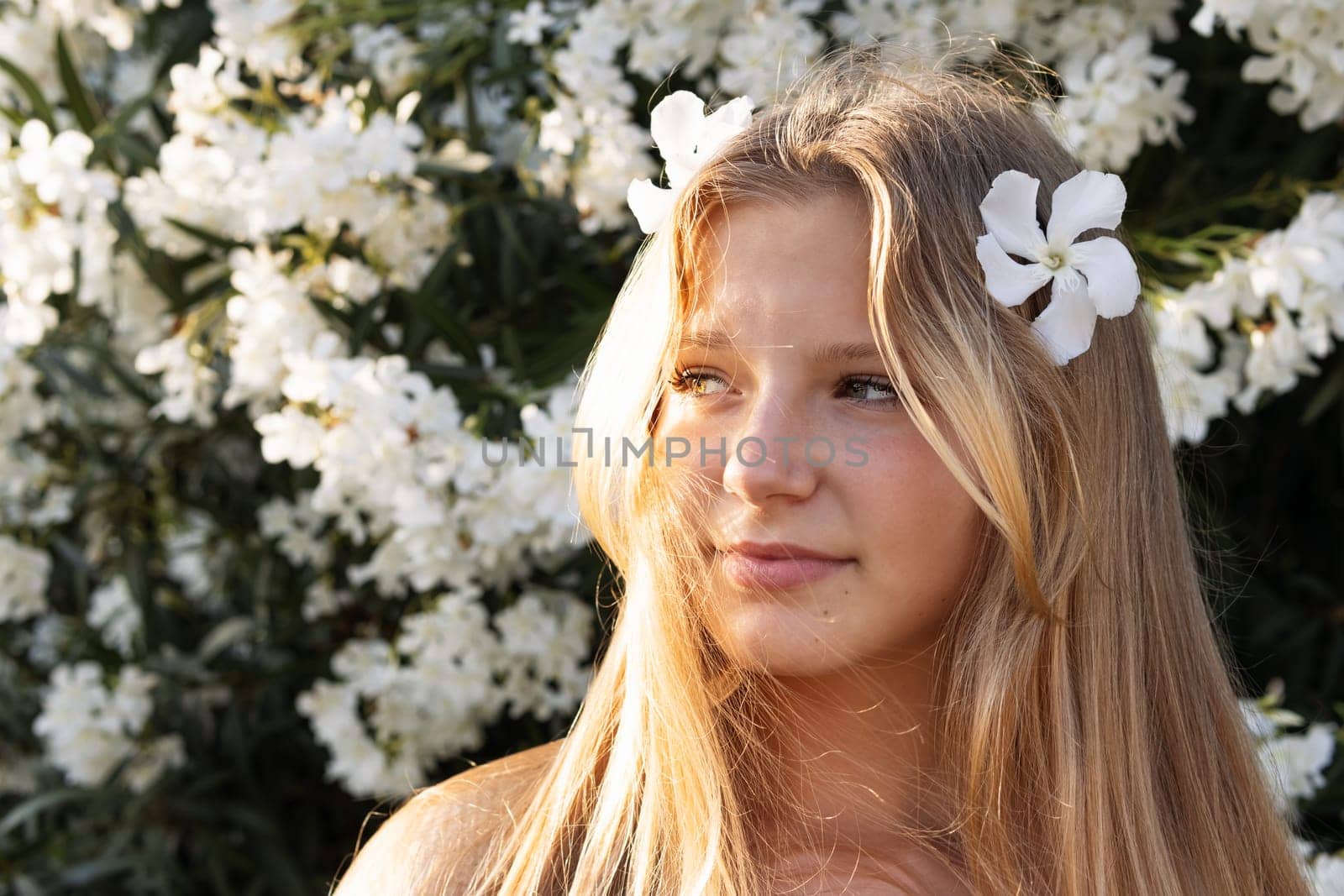 portrait of a teenage girl of European appearance, blond hair, the girl is standing on the street near a bush of white flowers, there is a place for an inscription. High quality photo