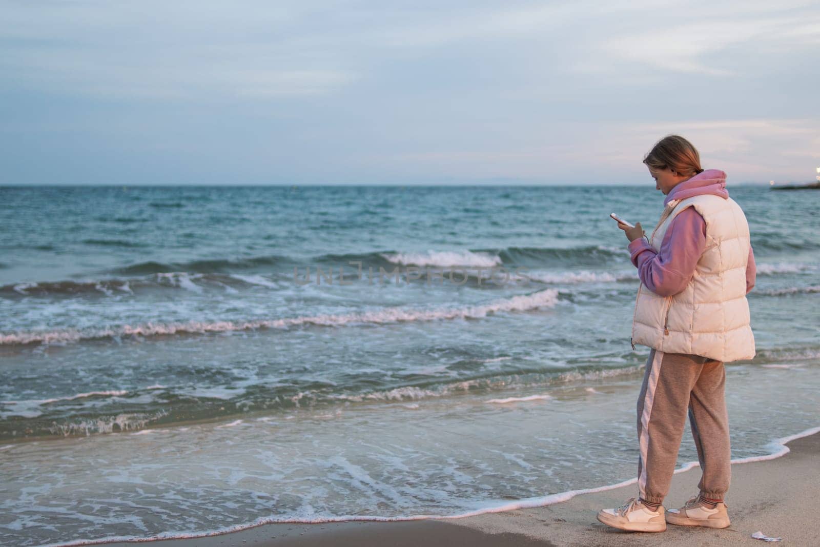 A girl of European make in casual clothes stands near the ocean with a phone in her hands is sad and looks into the phone. by PopOff