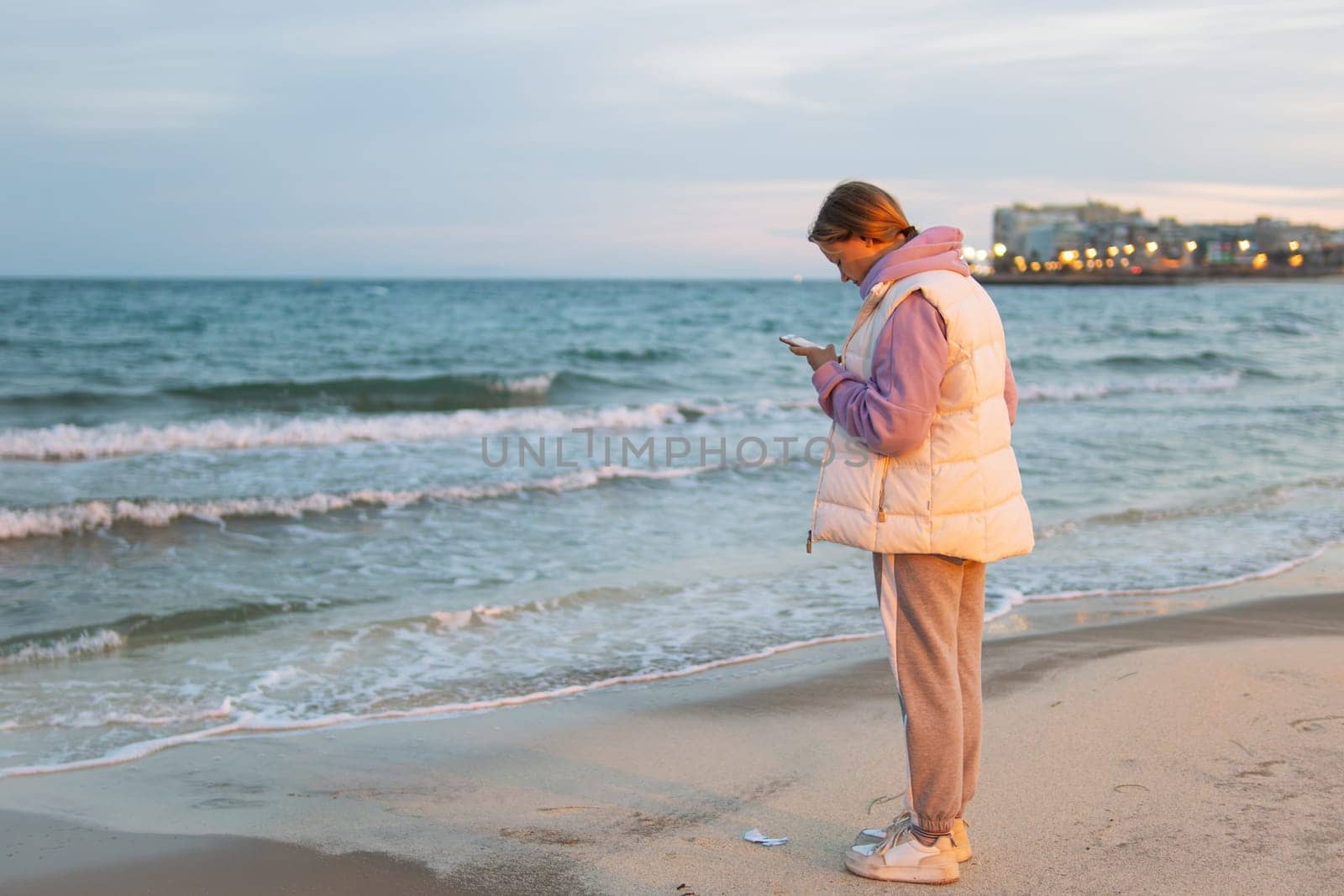 A girl of European make in casual clothes stands near the ocean with a phone in her hands is sad and looks into the phone. by PopOff