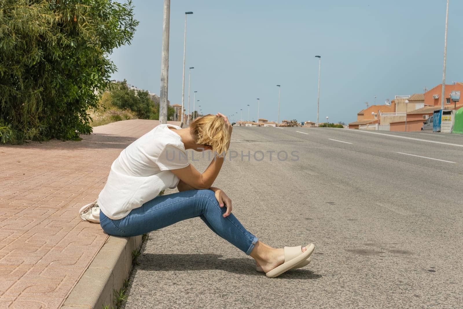 a girl sits on the pavement in jeans and a T-shirt, turned her back to the camera, looks at the road where cars drive. High quality photo