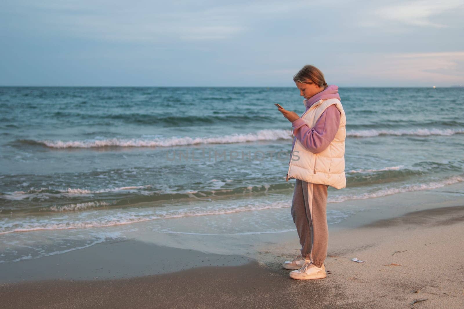 Portrait of an excited girl in her hands with a phone, standing on a beach with a blue sea. Sad and depressed young woman standing alone on the beach by the sea. Depression concept. High quality photo