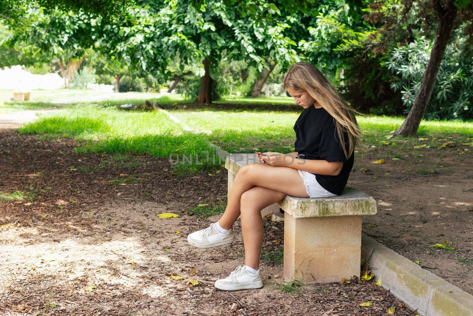 teenage girl in shorts and a black T-shirt sits on a park bench and looks at the phone. High quality photo