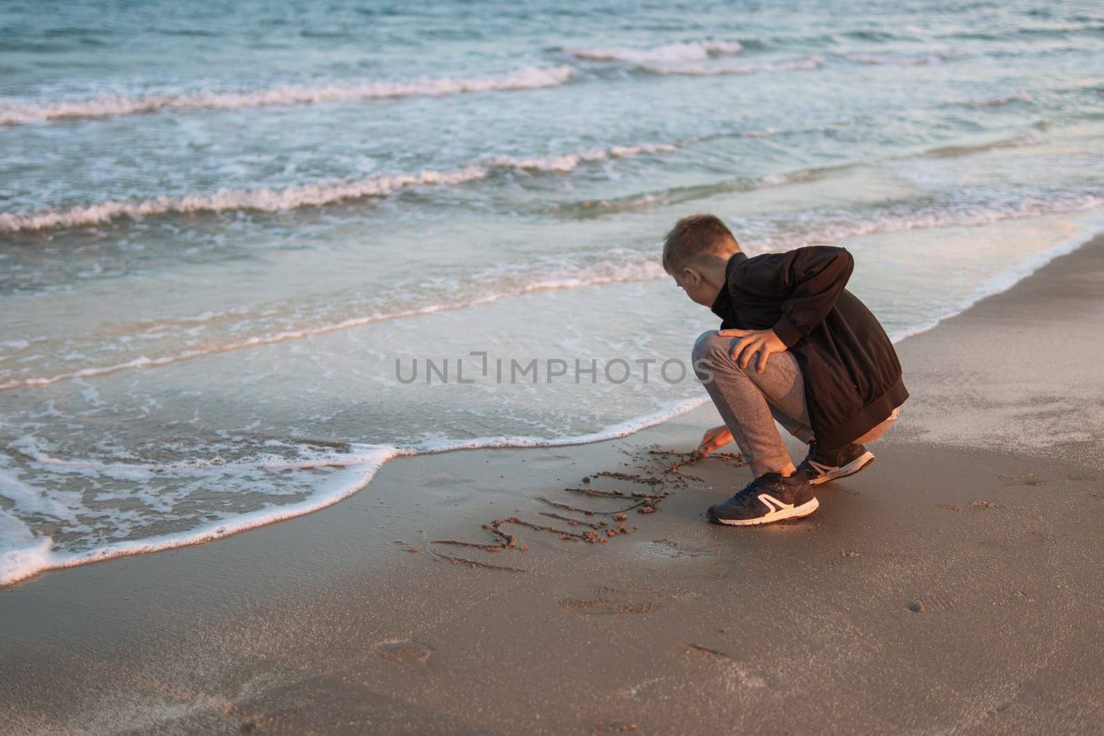 A boy of European appearance sits on the beach near the sea draws in the sand. A child on vacation sits near the sea by PopOff