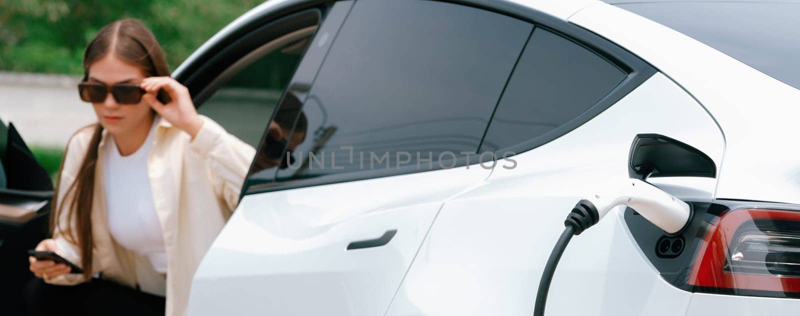 Young woman use smartphone to pay for electricity at public EV car charging station green city park. Modern environmental and sustainable urban lifestyle with EV vehicle. Expedient