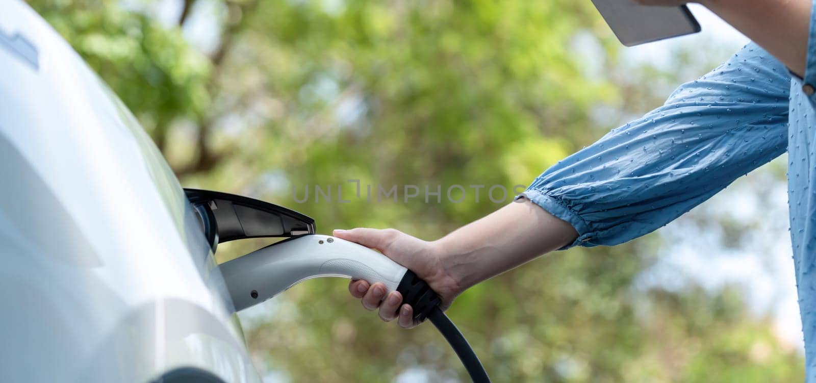 Panorama road trip vacation by the green countryside nature with beautiful young woman checking battery status from smartphone while recharging electric car. Eco-friendly travel wit EV car. Perpetual