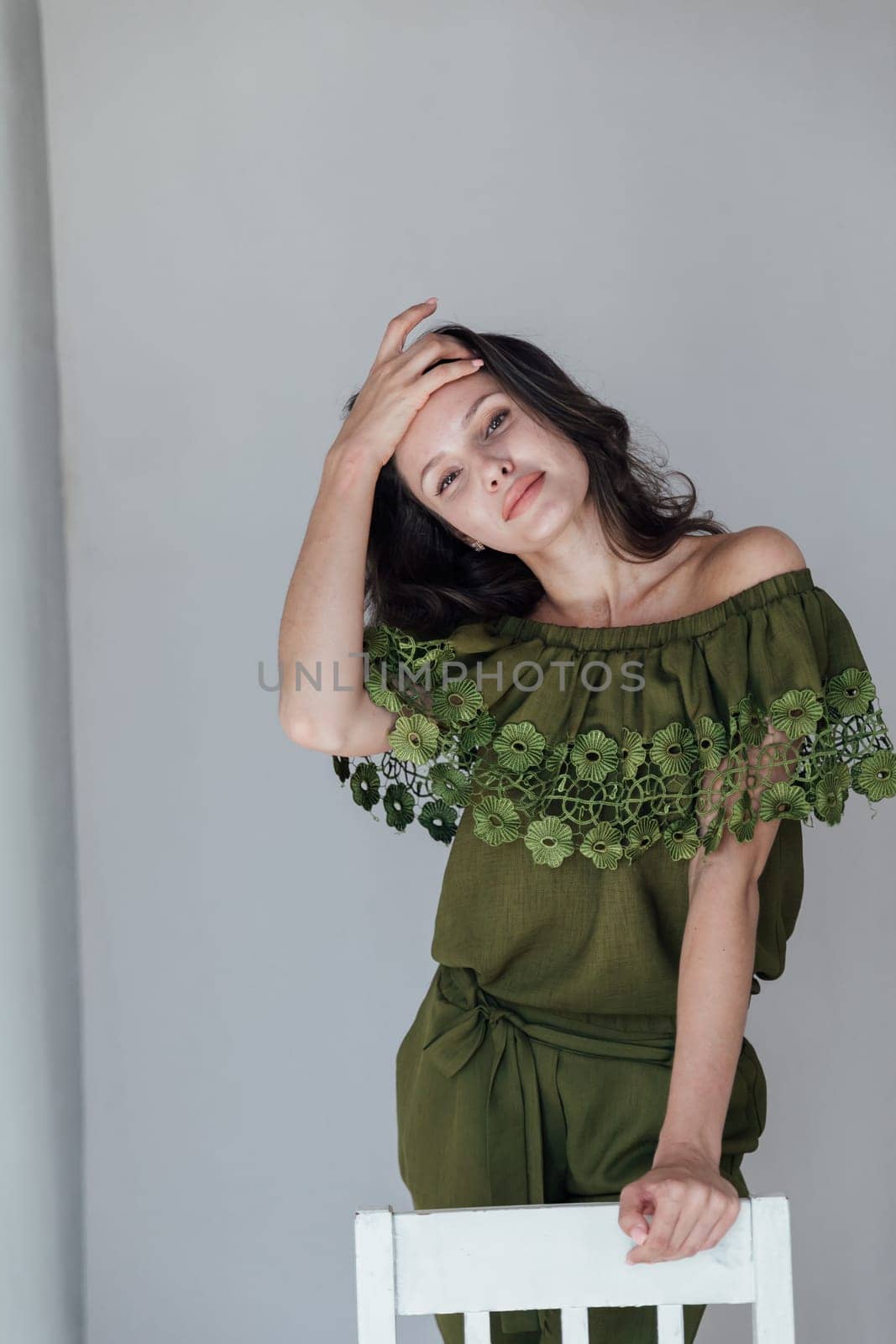 a brunette woman in green clothes adjusts her hair leaning on a white chair by Simakov