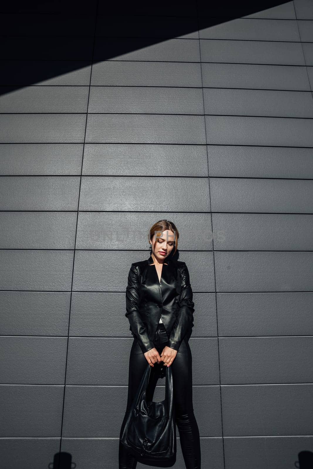 a fashionable woman in leather black clothes stands at a textured black wall by Simakov