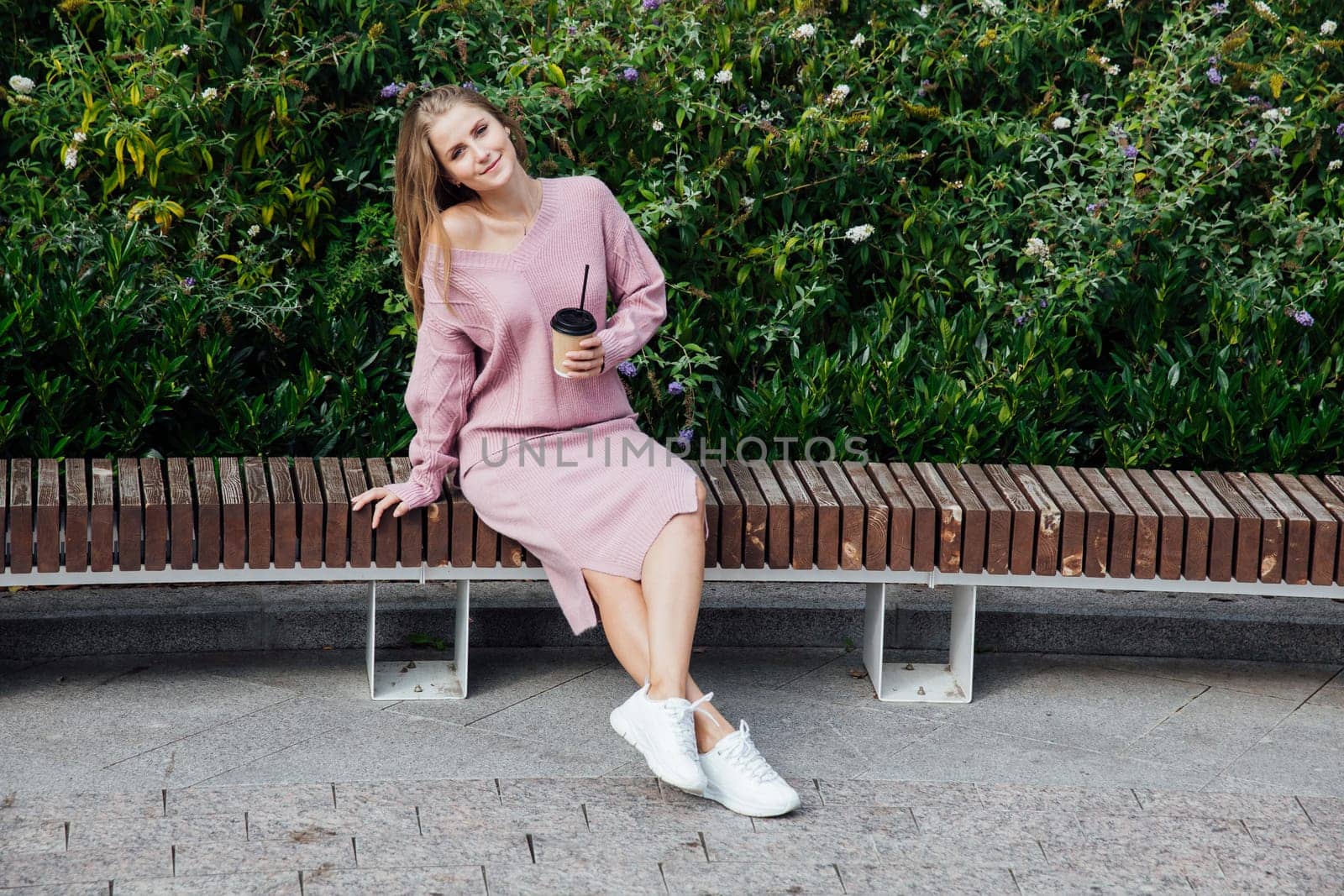 blonde woman sitting on bench with a cup of drink by the green bushes