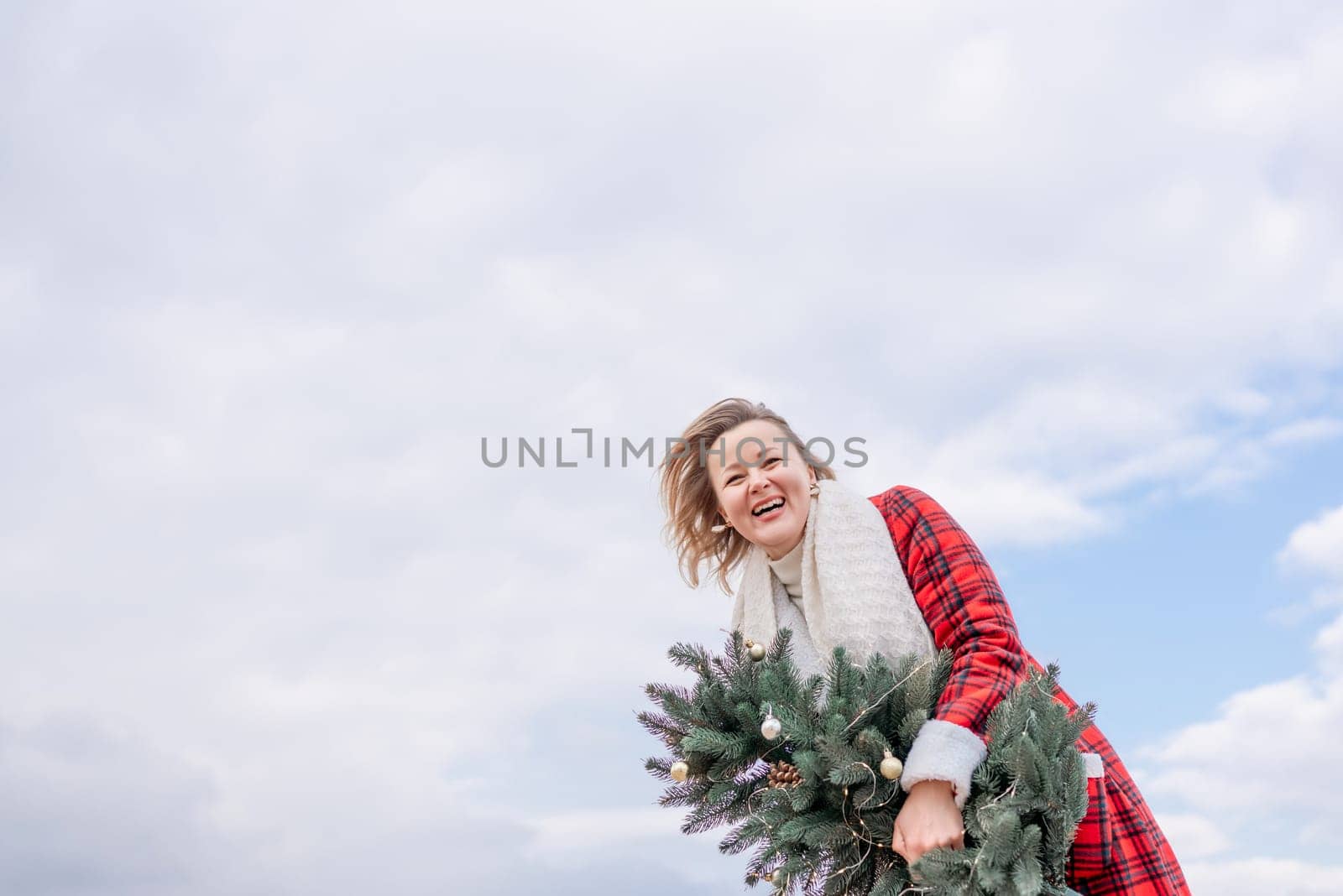 Blond woman holding Christmas tree by the sea. Christmas portrait of a happy woman walking along the beach and holding a Christmas tree in her hands. Dressed in a red coat, white suit
