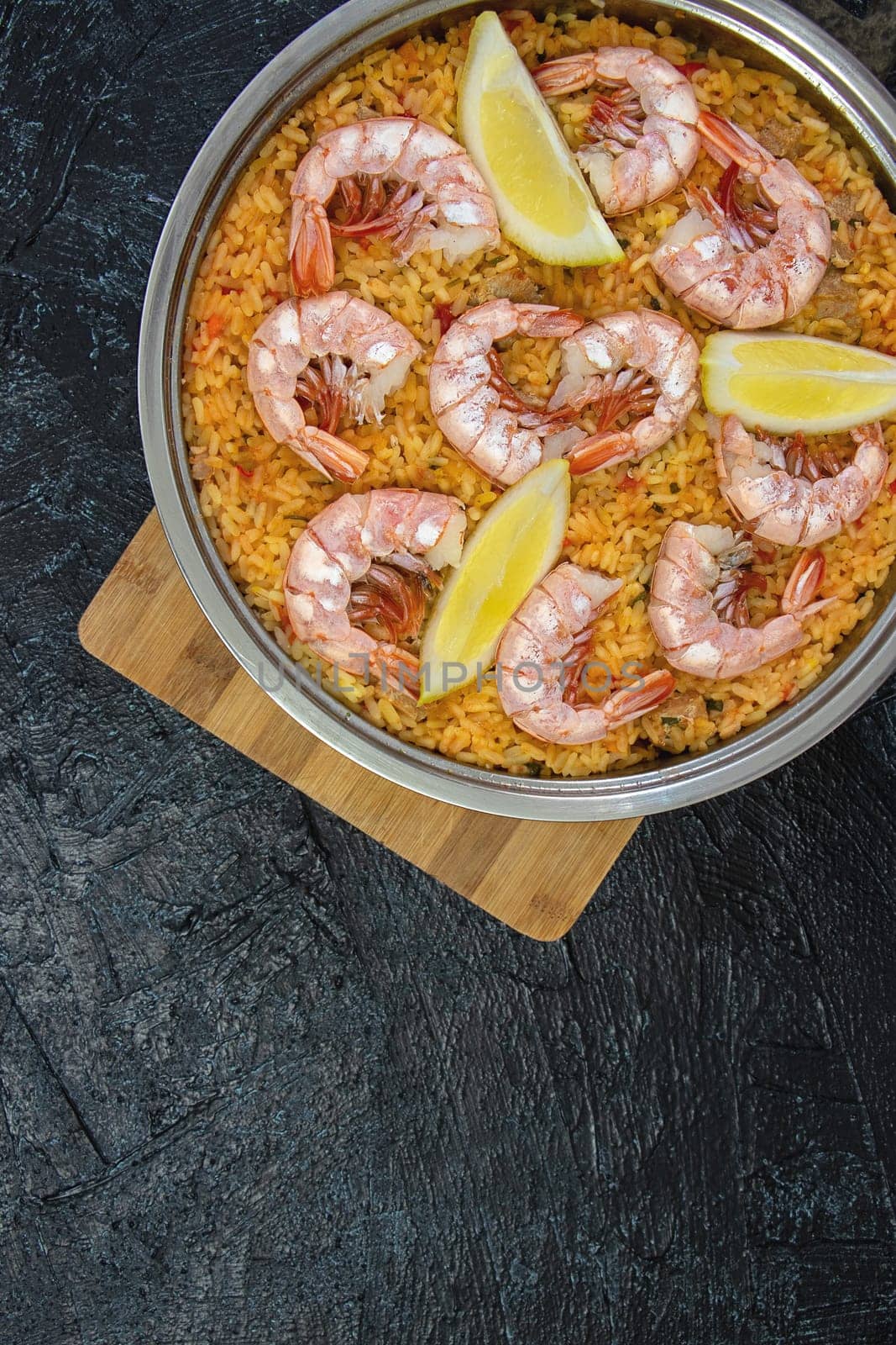 Vertical photo with top view of frying pan with paella on wooden board on a black textured background. flat lay pan with rice, shrimp, lemon wedges, saffron, copy space. soft focus. by Leoschka