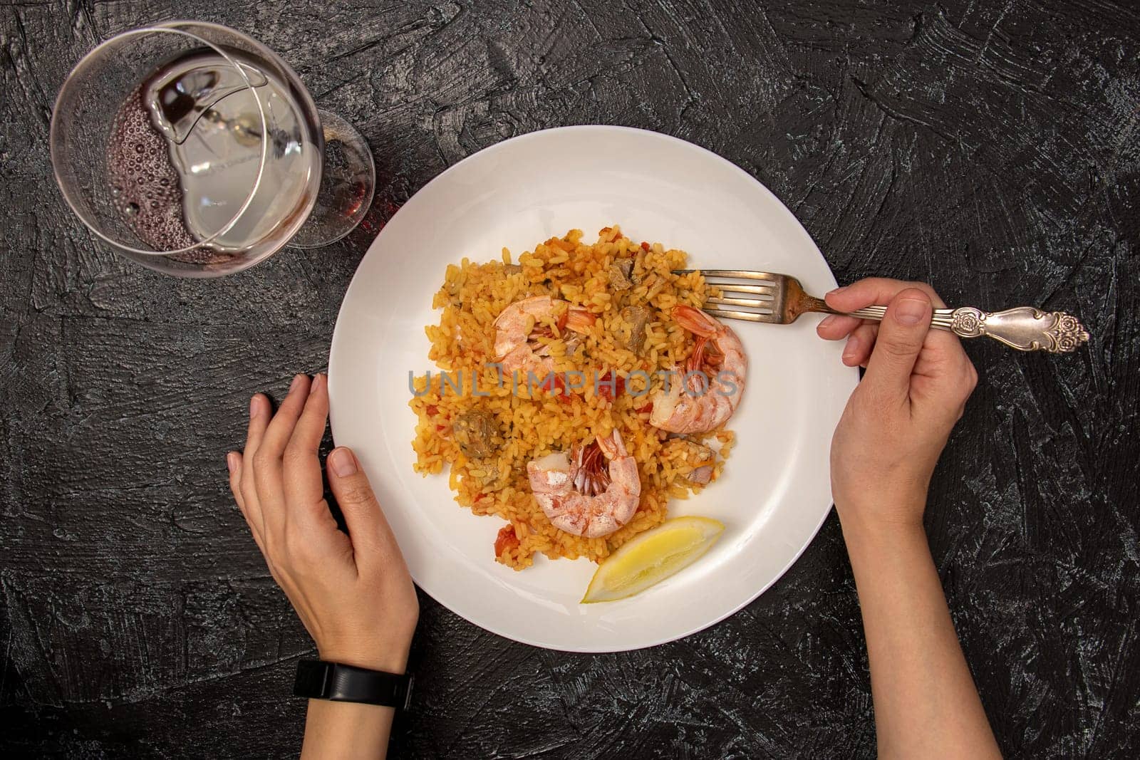 top view of female hands with fork, white plate with paella and shrimp and glass of red wine on black textured background. dinner alone. by Leoschka