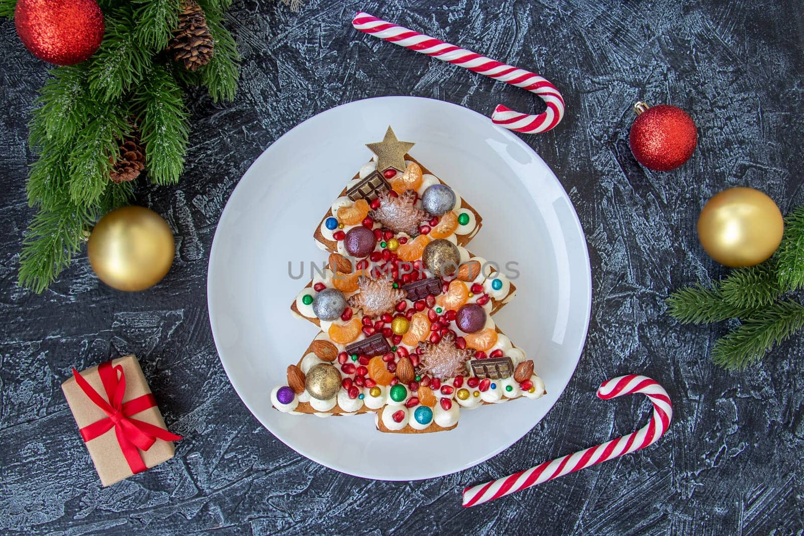 flat lay of christmas cake on black textured background with christmas balls and candy sticks. top view of christmas dessert with chocolate, pomegranate seeds, tangerines and nuts