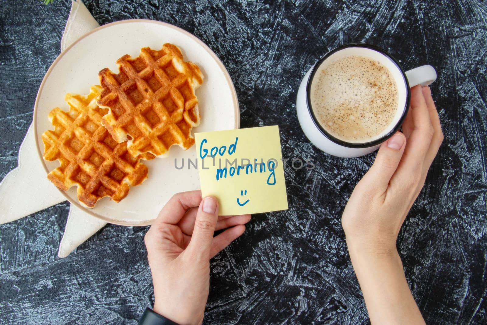 Top view of womans hands holding a note with words GOOD MORNING next to cup of coffee and spotted homemade Viennese waffles on black textured background. breakfast flat lay by Leoschka
