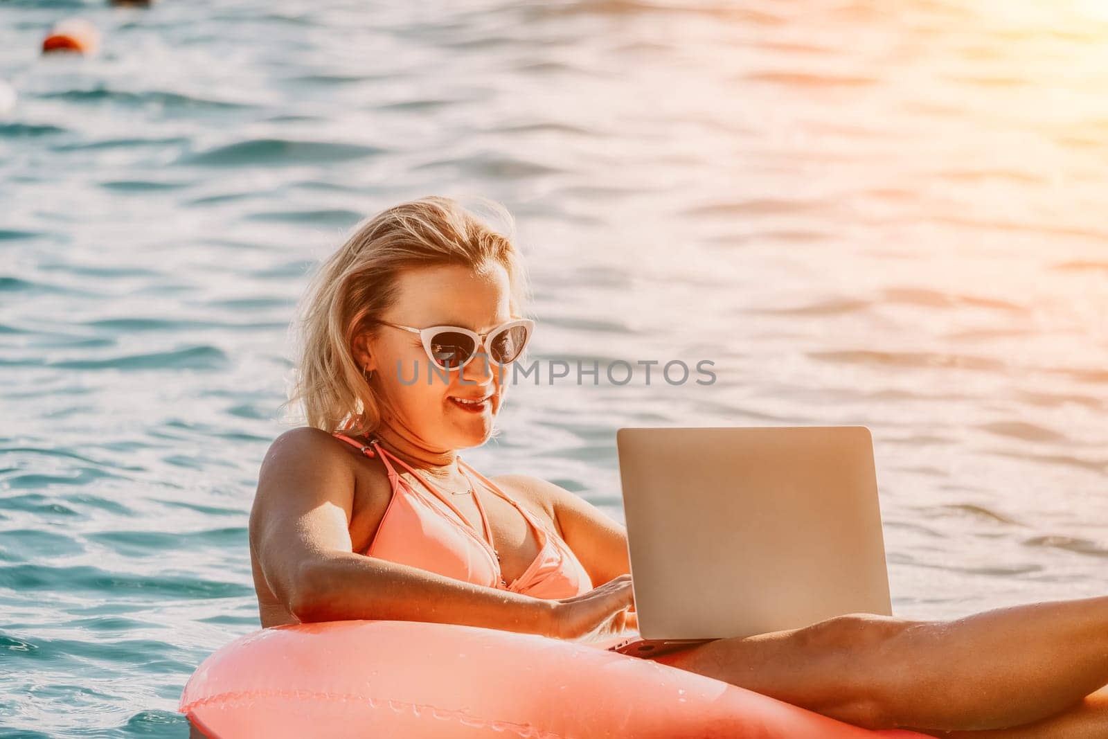 Woman works on laptop in sea. Freelancer, young blond woman in sunglases floating on an inflatable big pink donut with a laptop in the sea at sunset. Freelance, travel and holidays concept. by panophotograph