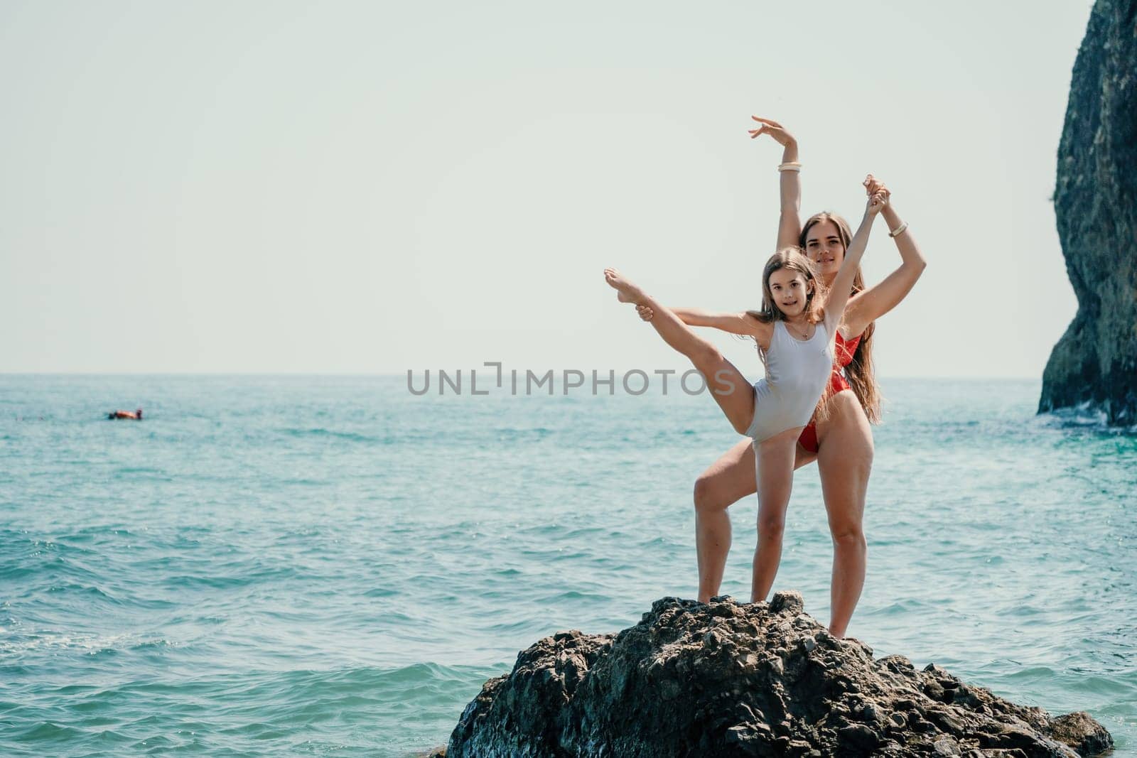 Woman and her daughter practicing balancing yoga pose on one leg up together on rock in the sea. Silhouette mother and daughter doing yoga at beach by panophotograph