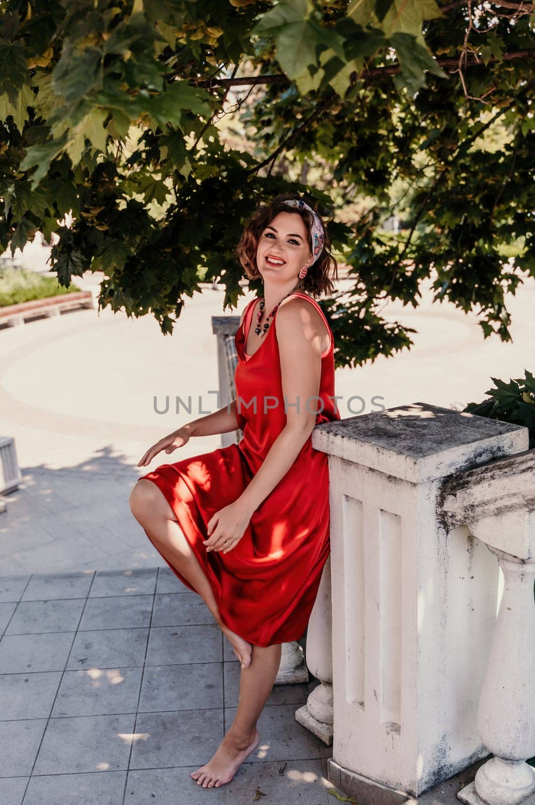 A pretty woman in a red silk dress and a bandage on her head smiles against the background of the leaves of a tree. She is leaning on the coop and looking into the camera. Vertical photo. by Matiunina