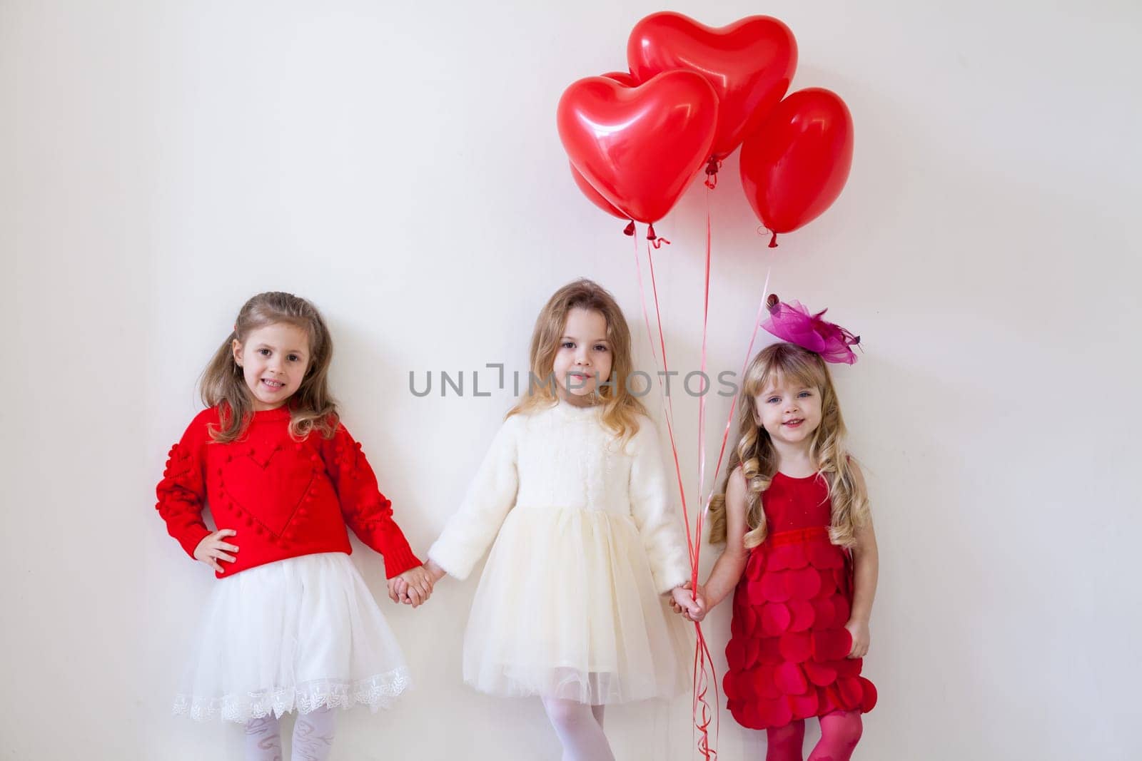 Three elegant little girls stand with red valentine's day holiday balls beautifully dressed