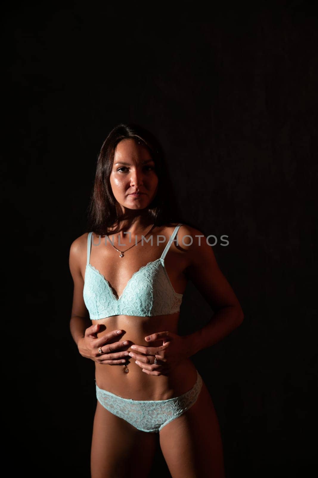 a brunette woman in blue lingerie stands holding on to her stomach on a black background