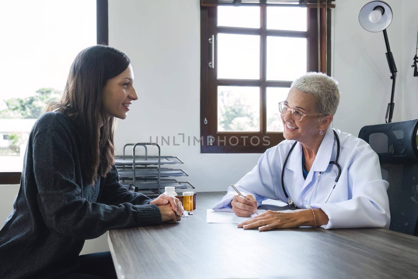 Elderly female doctor in white coat talking with beautiful young patient in clinic, giving advice on heart disease treatment and health care and medicine, medicine concept by wichayada