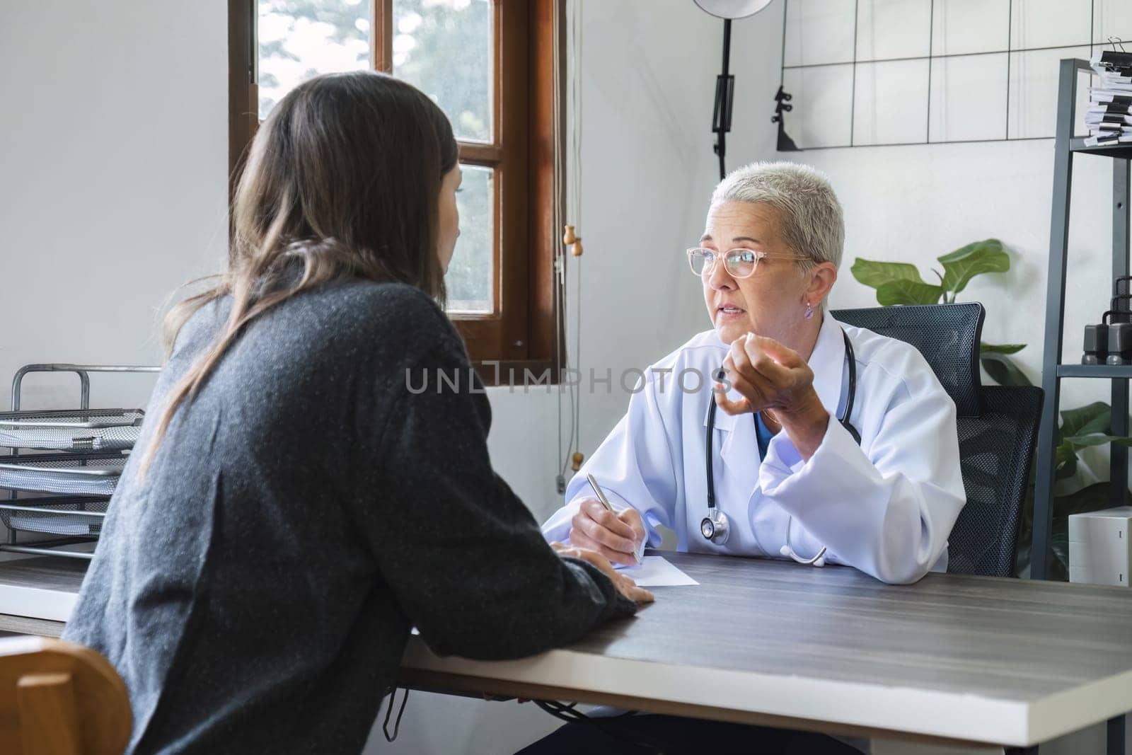 Elderly female doctor in white coat talking with beautiful young patient in clinic, giving advice on heart disease treatment and health care and medicine, medicine concept by wichayada