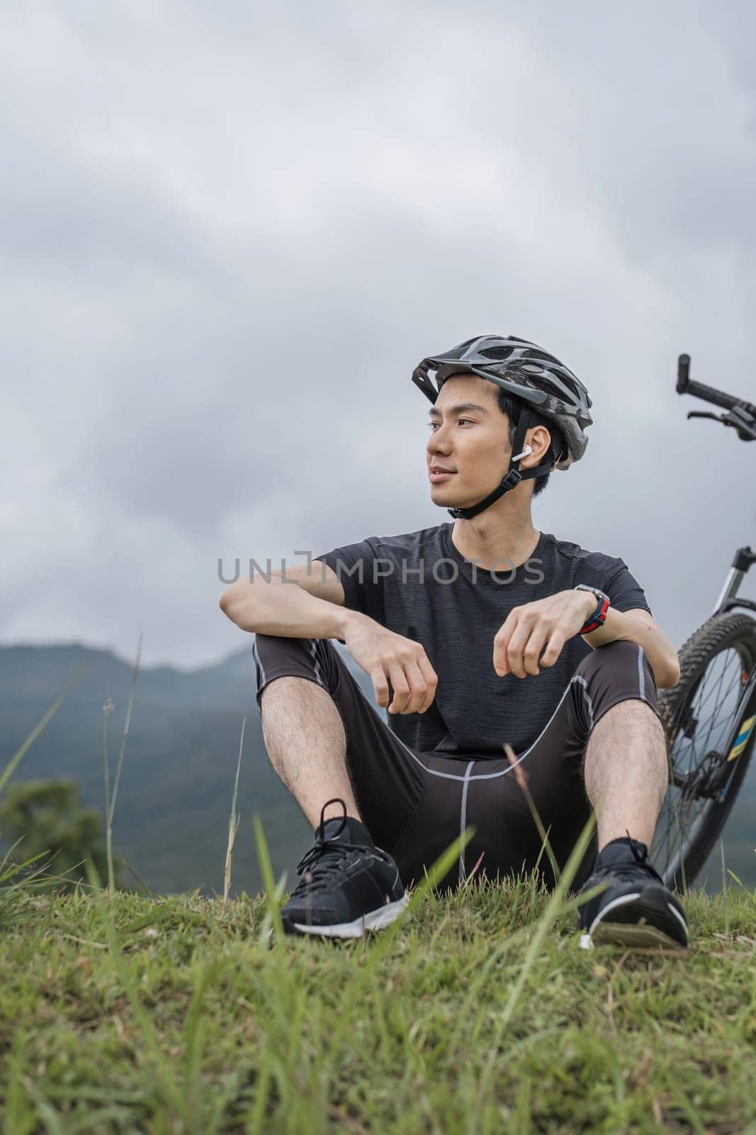 A happy and handsome Asian man in sportswear and a bike helmet enjoys listening to music through his earbuds while riding a bike on the country roads on the weekend. by wichayada