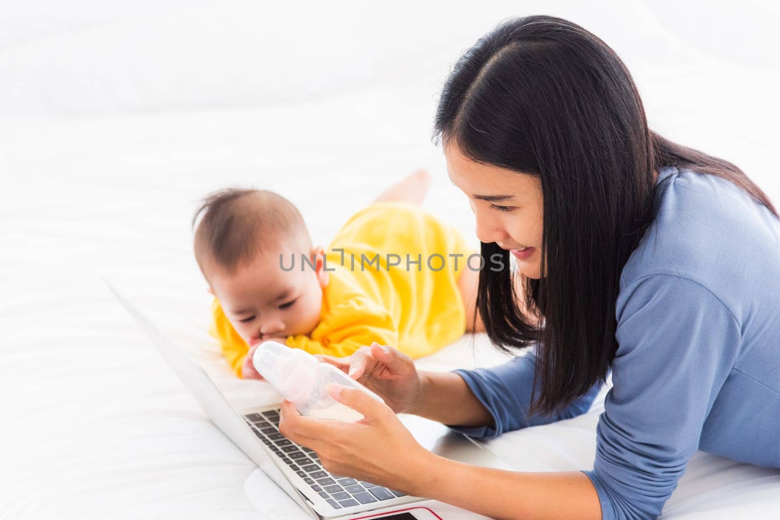 Asian new young beautiful mother finding and reads information on a laptop for milk mixing on a bottle in the bedroom at home