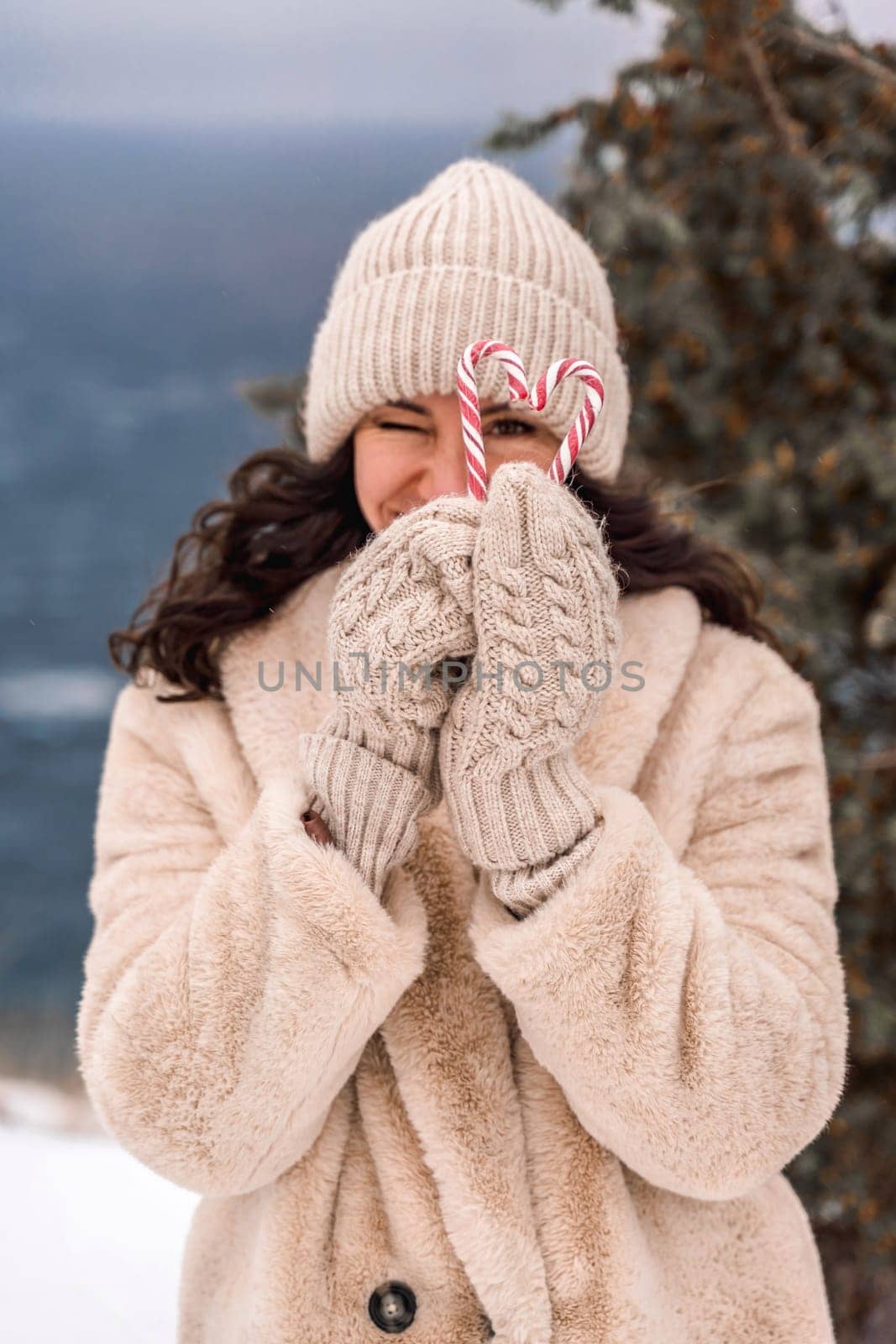 Woman christmas candy. Smiling woman in knitted hat, mittens and beige coat holding lollipops christmas candy canes in her hands in shape of heart against the backdrop of the sea. by Matiunina