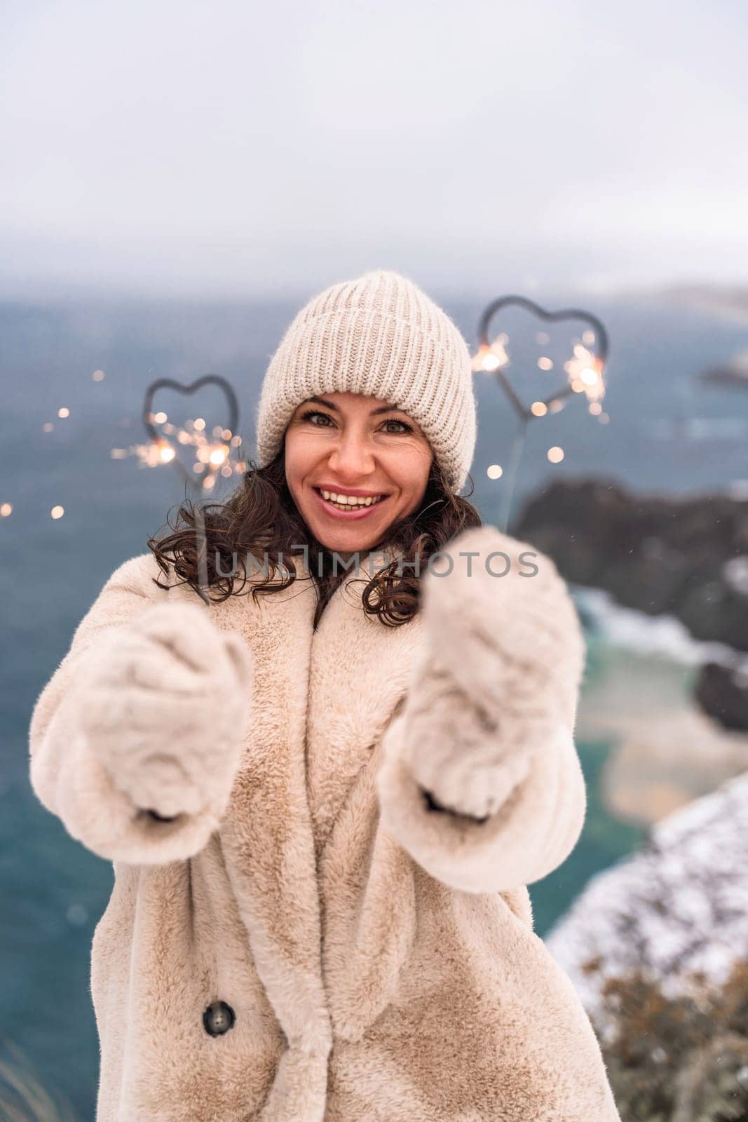 Outdoor winter portrait of elegant happy smiling beige hat, light faux fur coat holding heart sparkler, posing against sea and snow background