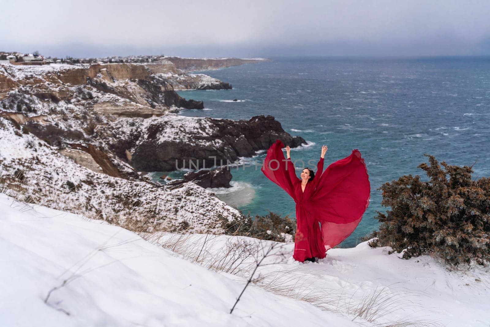 Woman red dress snow sea. Happy woman in a red dress in the snowy mountains by the emerald sea. The wind blows her clothes, posing against sea and snow background