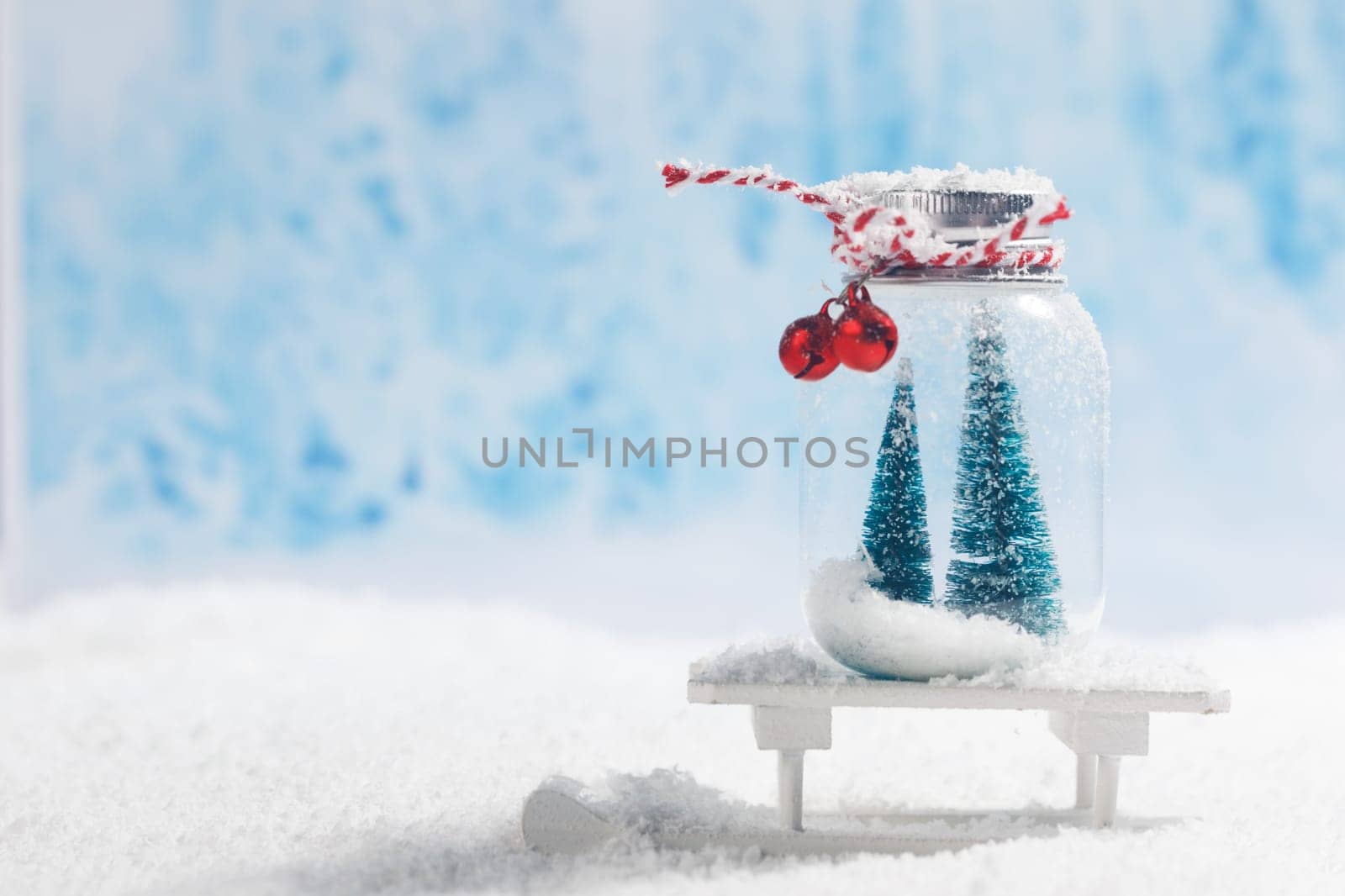 Christmas ornaments of glass jar with striped ribbon , bells and fir trees inside on toy sled on snow forest background