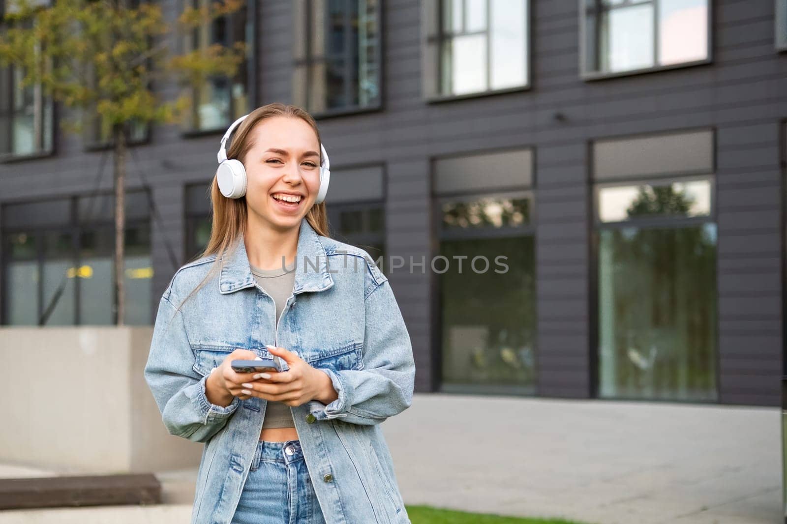 A young woman in headphones and smiling her gaze towards the camera, holding a phone in hand.