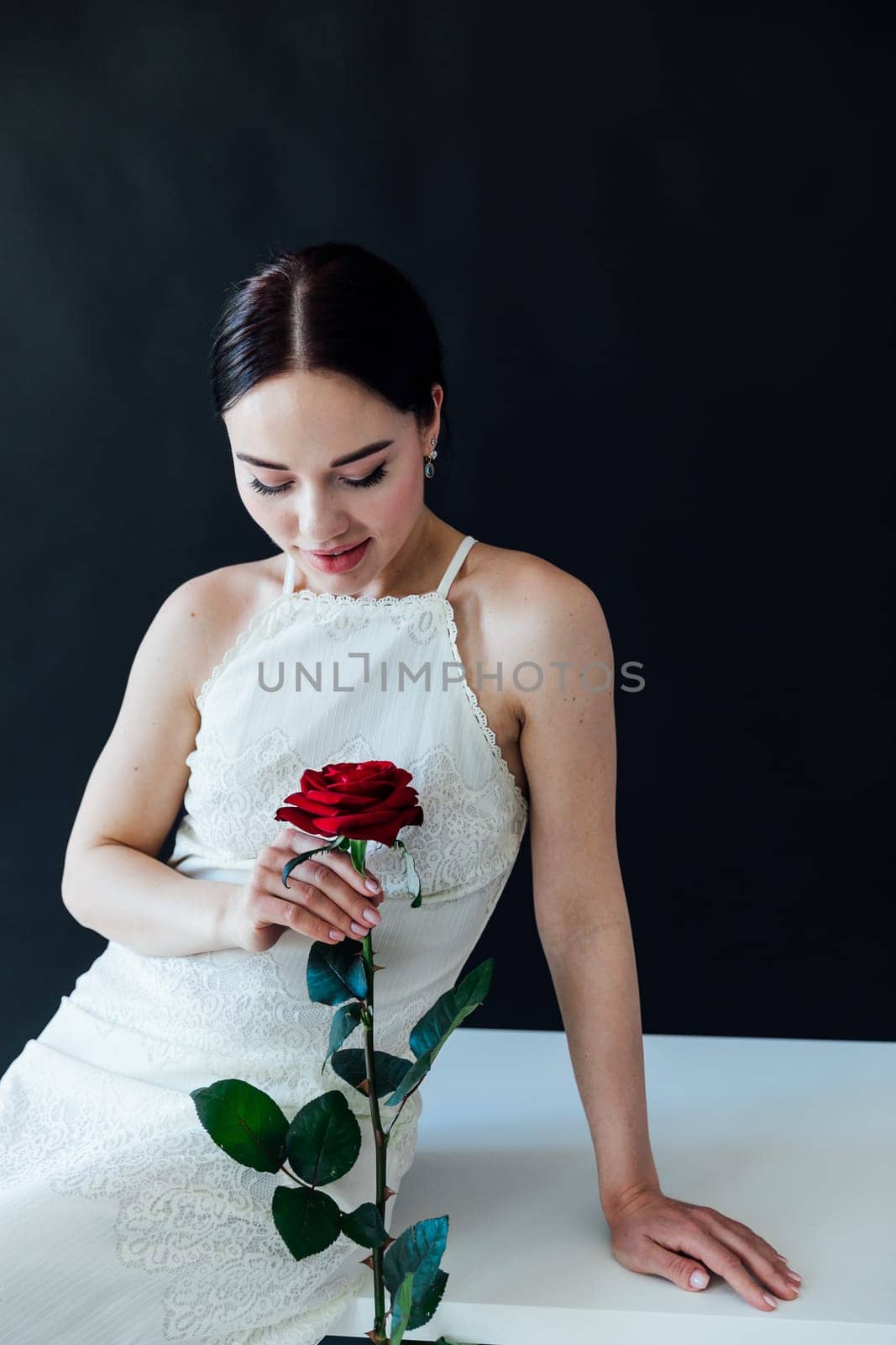 beautiful brunette in white dress with a red rose sits on a table against a black wall