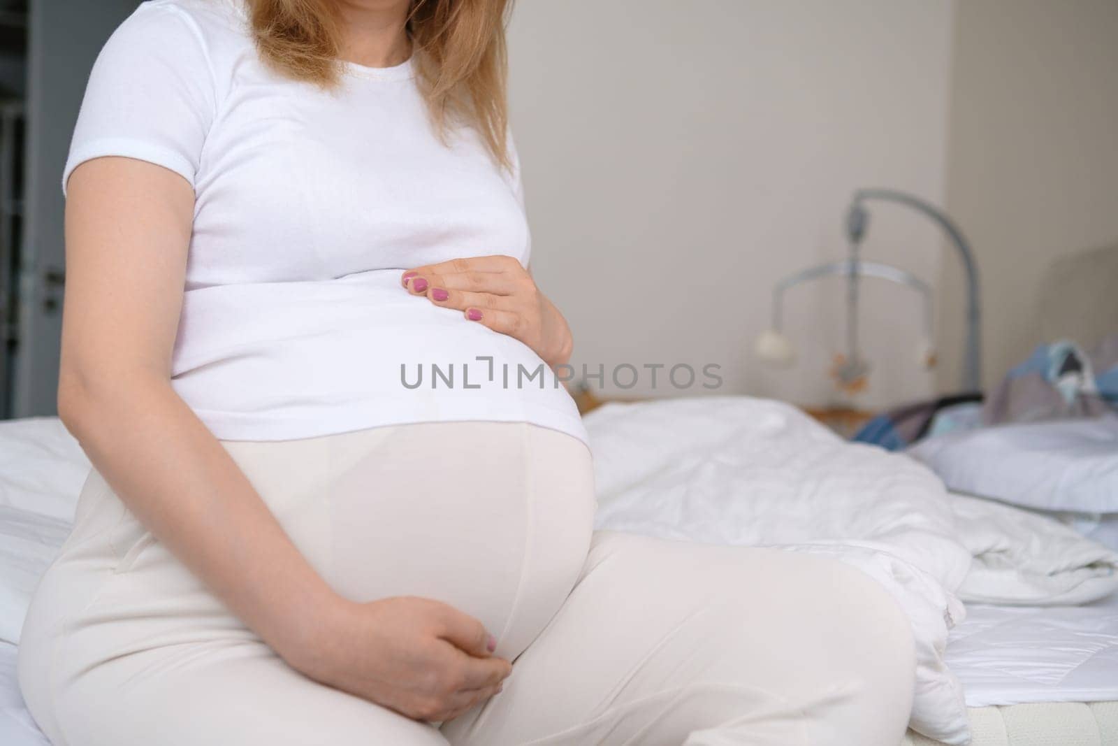 Pregnant woman sits on the bed with a baby crib adorned with hanging toys on the background.