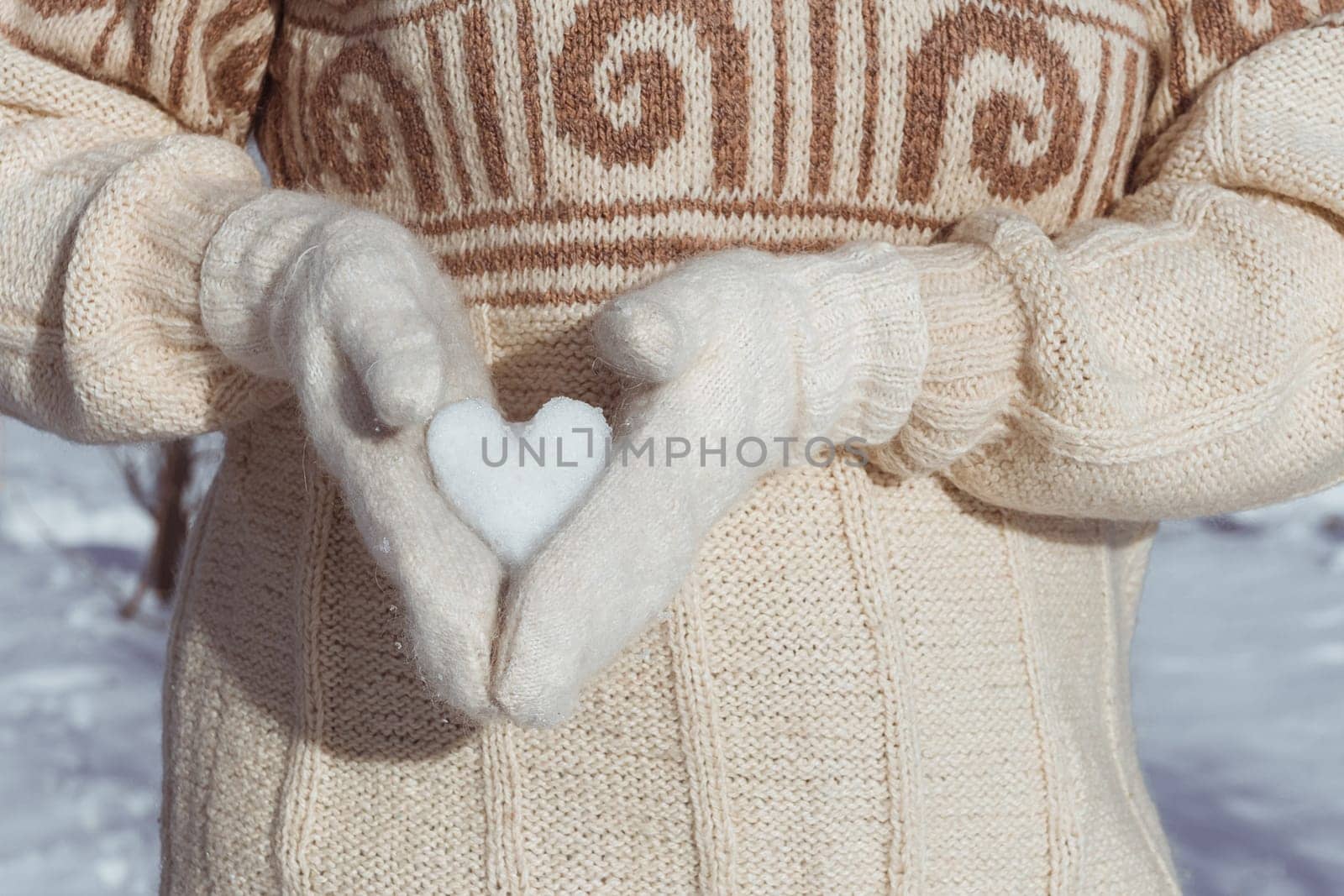 A girl in a beige sweater holds a heart made of snow in her hands, dressed in white mittens, from the side. A gift for Valentine's Day. Postcard with copy space by ElenaNEL