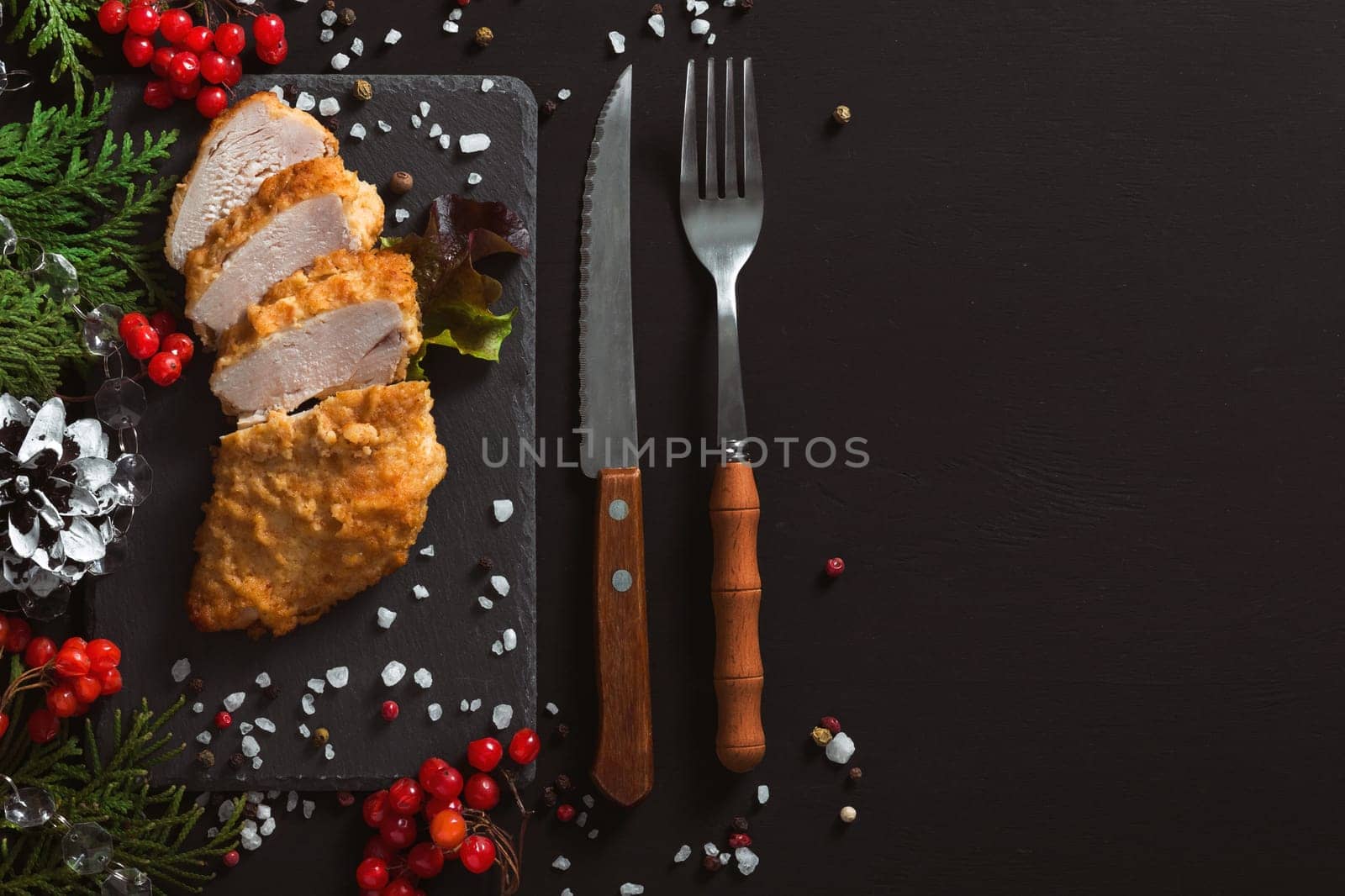 White chicken meat fried in batter on a black background with Christmas decorations and cutlery. The chicken breast is cut into pieces. Top view, copy space