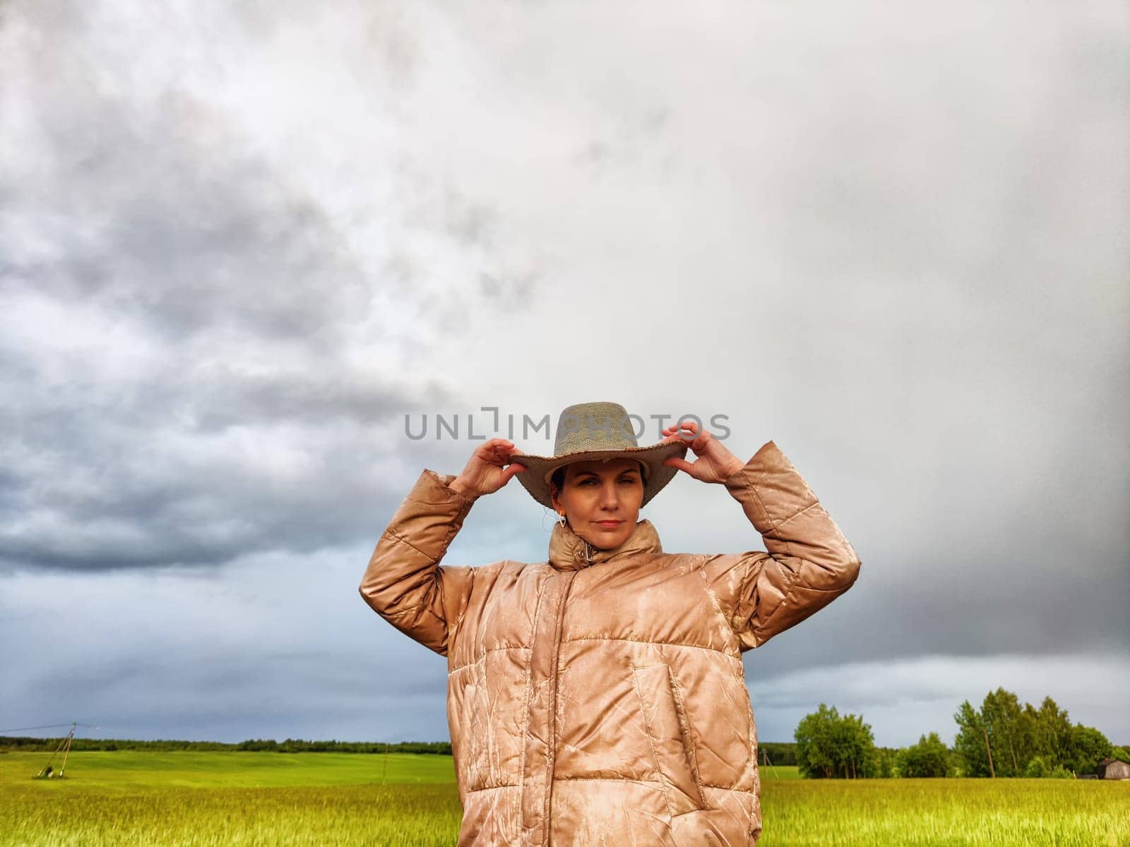 An adult girl looking like a cowboy in a hat in a field and with a stormy sky with clouds takes pictures of rainbow and takes selfie in the rain. Woman having fun outdoors on rural and rustic nature