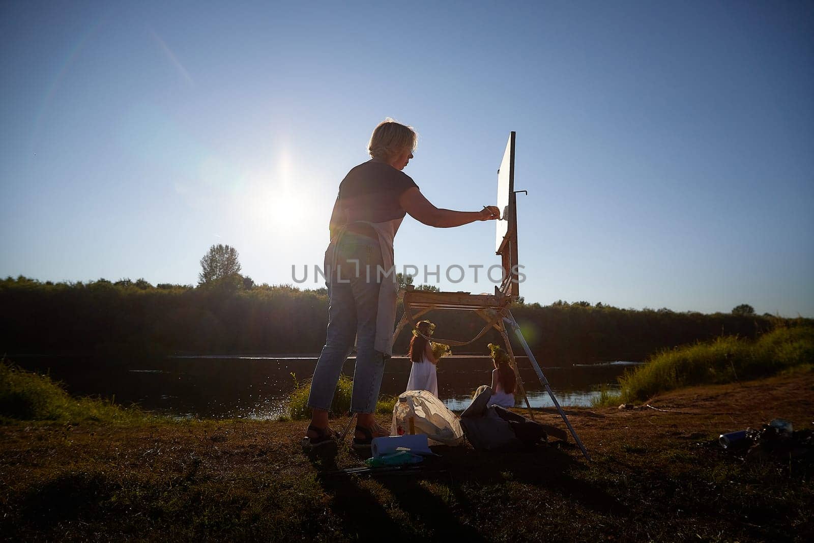 Adult female artist painting picture near water of river or lake in nature and girls in white sundress and flower wreath posing in the holiday of Ivan Kupala in nature at sunset by keleny