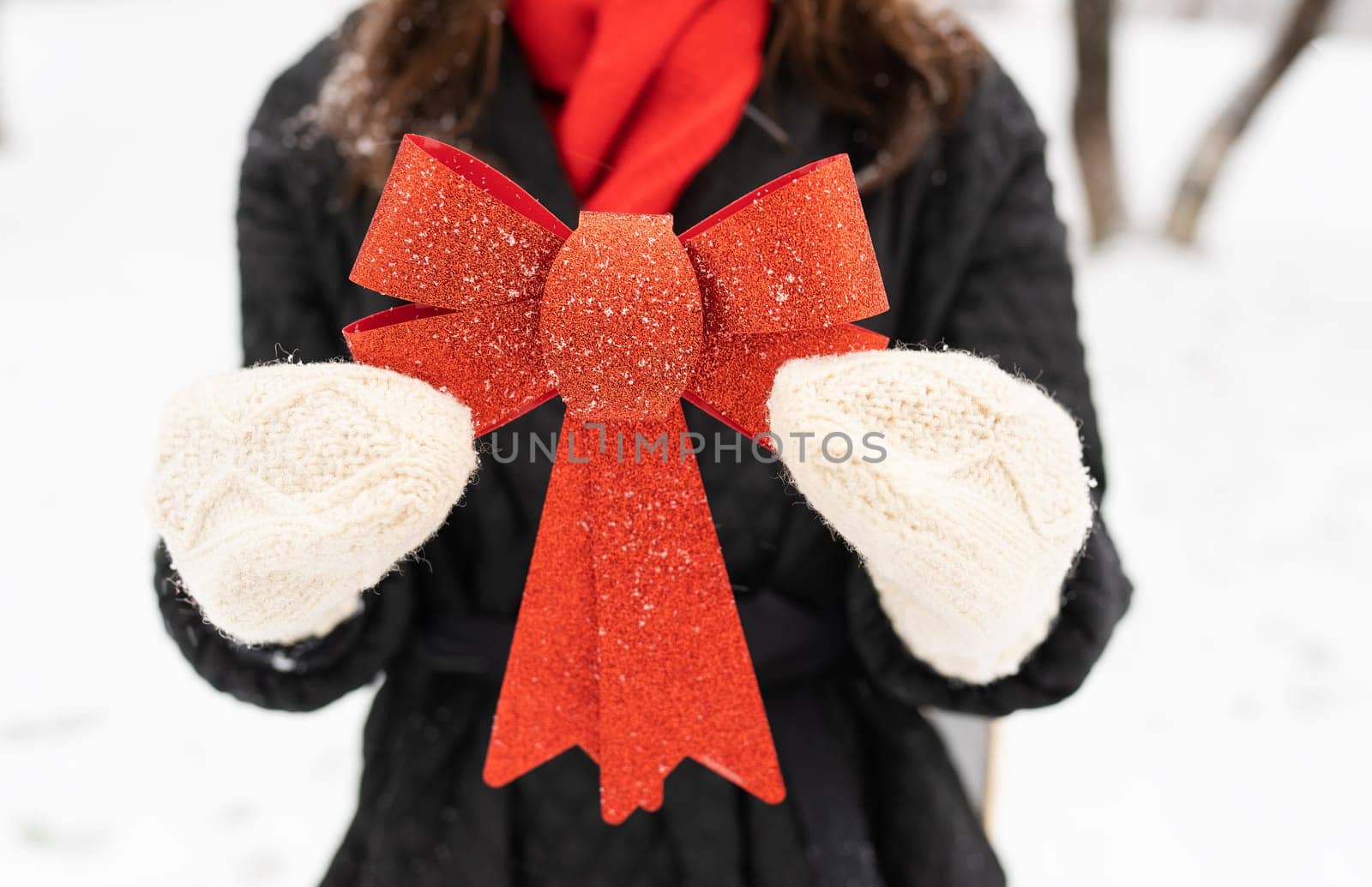 A woman in a black coat and red scarf holds a large red shiny bow in a snowy winter scene. Christmas and New Year concept