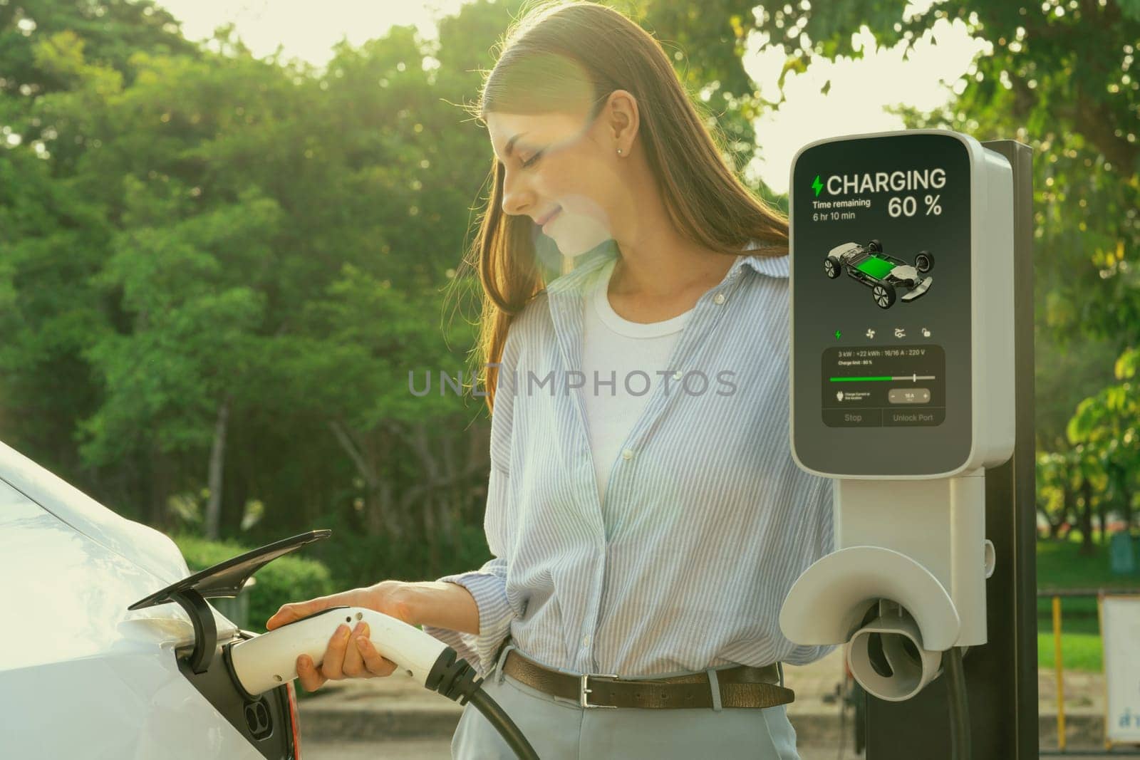 Young woman recharging battery for electric car during road trip travel EV car in natural forest or national park with sunrise lighting. Eco friendly travel during vacation and holiday. Exalt