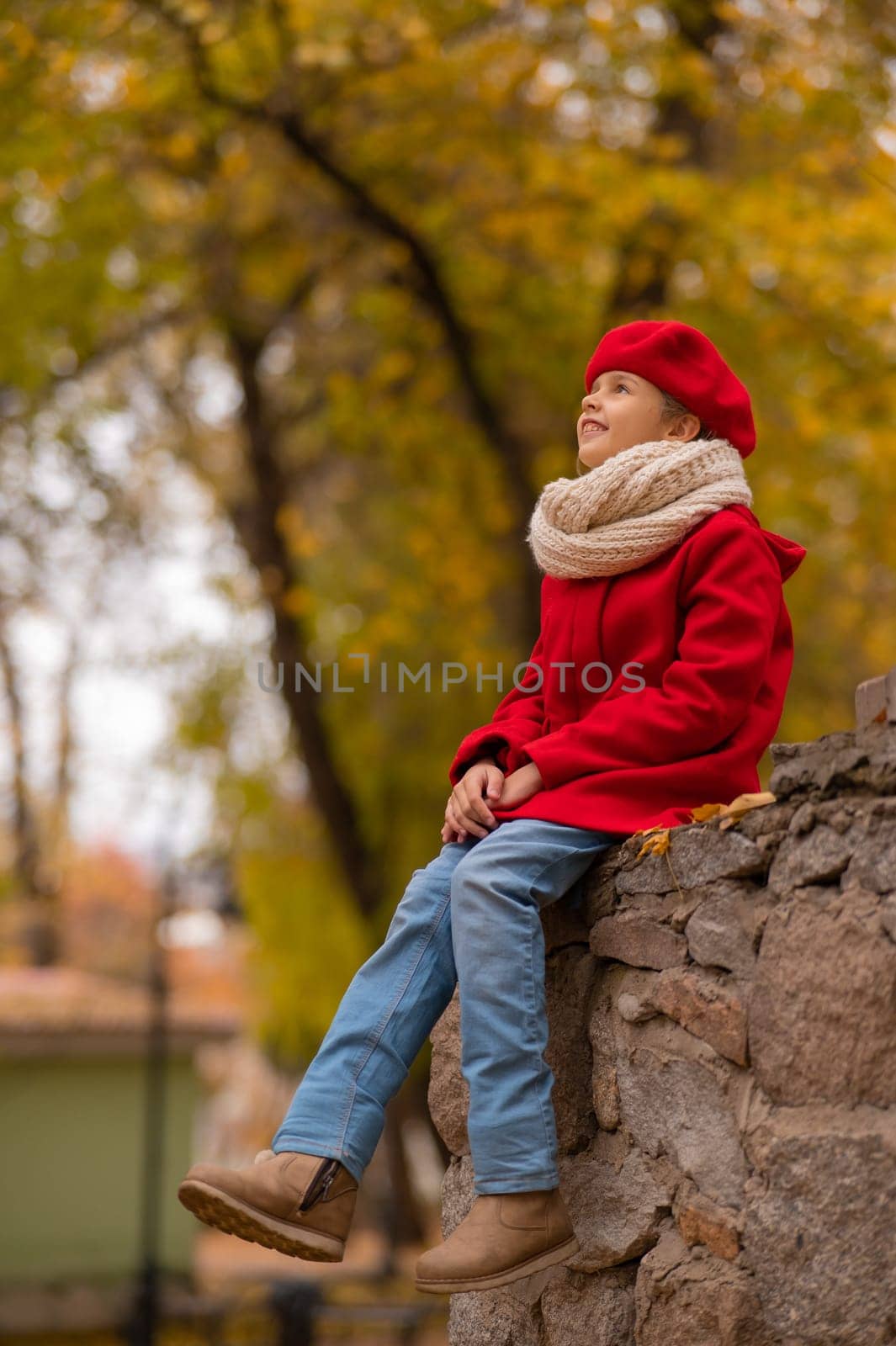 Smiling caucasian girl in a red coat and beret sits on a brick wall on a walk in autumn