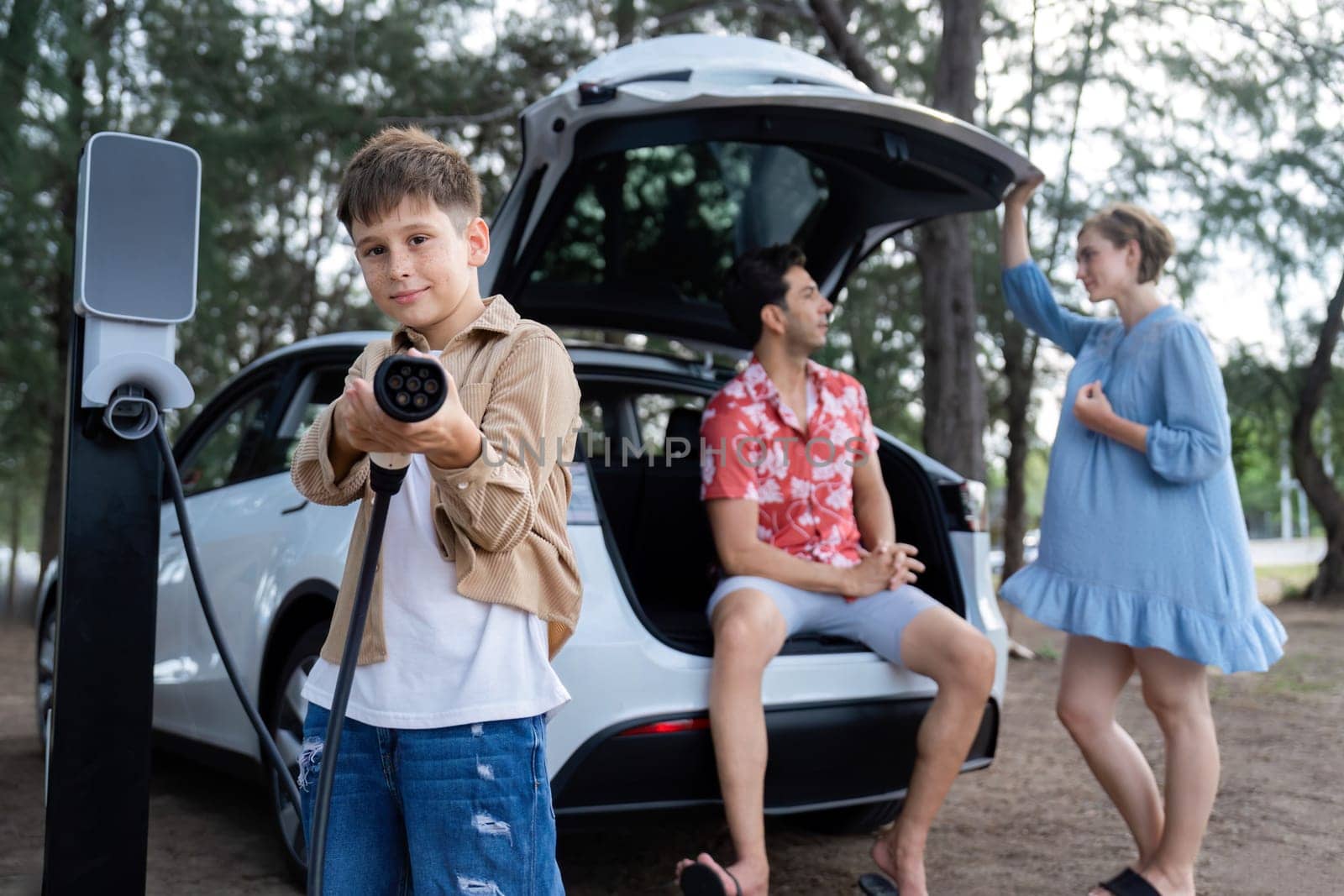 Little boy holding EV charger and point at camera with his family sitting on the trunk in background. Road trip travel with alternative energy charging station for eco-friendly car concept. Perpetual