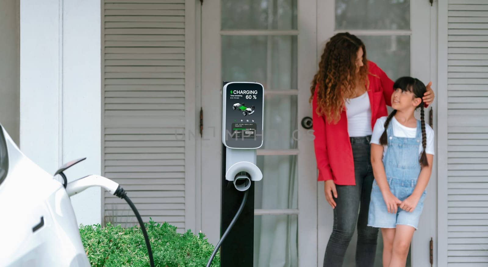 Happy little young girl learn about eco-friendly and energy sustainability as she help her mother recharge electric vehicle from home EV charging station. EV car and modern family. Panorama Synchronos