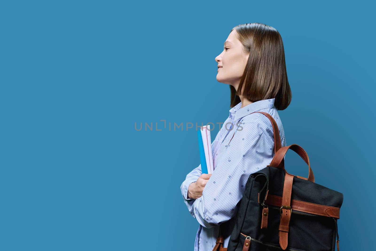 Profile view of young woman college student with backpack textbooks on blue studio background, copy space space for advertising image text. Beauty fashion care cosmetics face skin hair