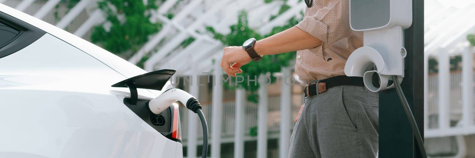 Young man checking time on smartwatch while EV charger to recharging battery from charging station in parking lot. Rechargeable EV car for sustainable eco friendly urban travel. Panorama Expedient